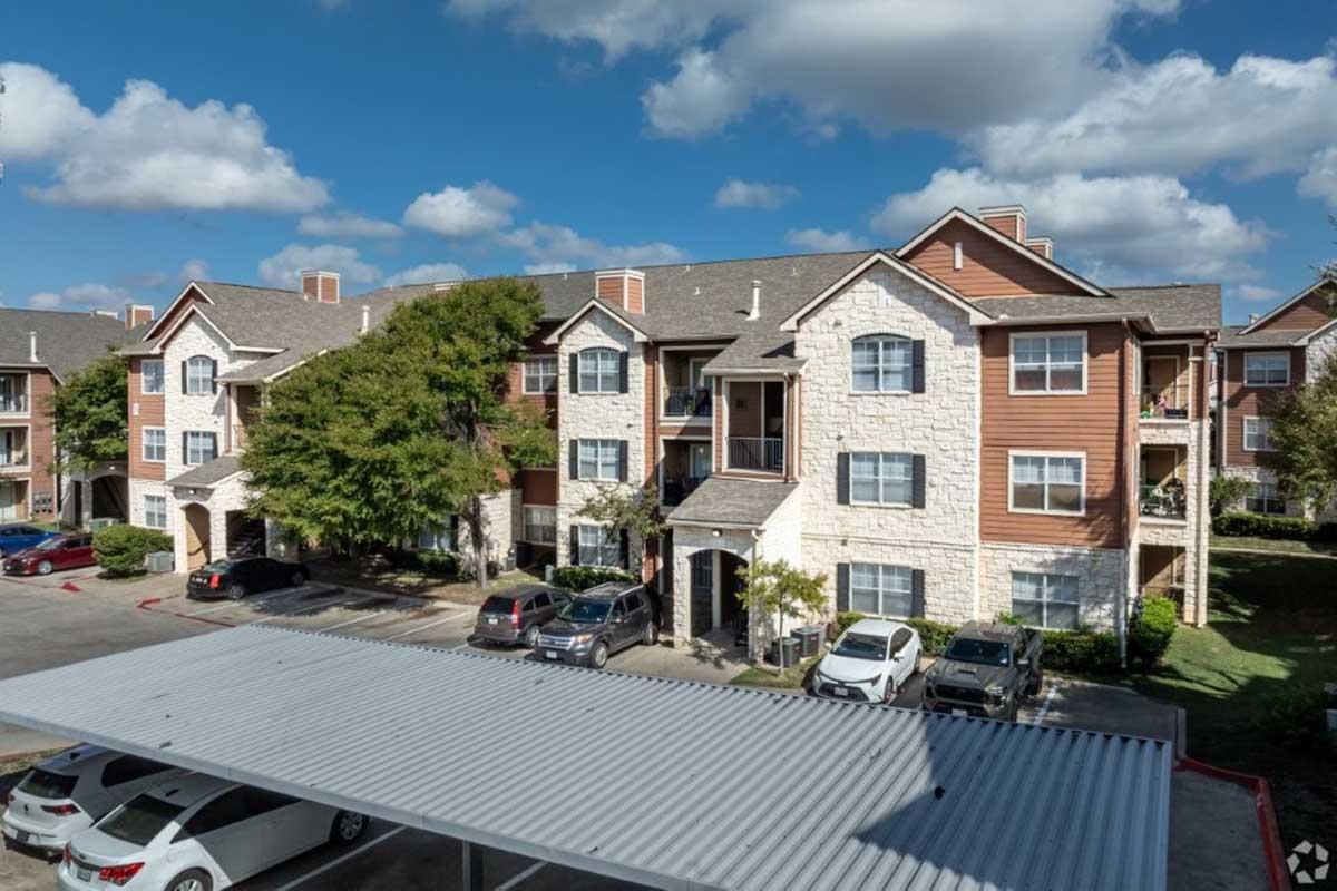 Exterior view of a modern apartment complex featuring multiple buildings with a mix of stone and wood siding. The scene includes a parking area with several cars and a canopy. The sky is partly cloudy, adding to the pleasant setting. Lush greenery surrounds the buildings, creating a welcoming atmosphere.