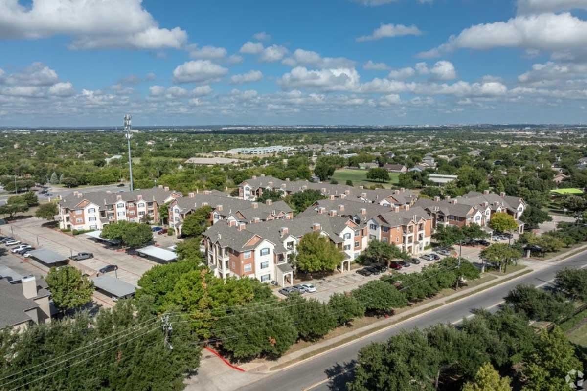 Aerial view of a residential complex with multi-story buildings surrounded by trees and parked cars. There are green fields and a clear blue sky with clouds in the background, showcasing a suburban landscape.