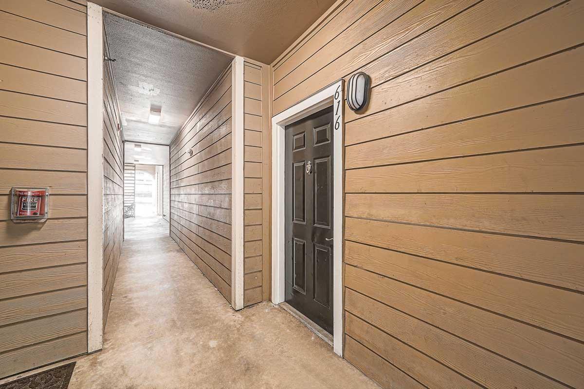 A hallway in a building featuring brown wooden panel walls. On the left, there is a mailbox mounted on the wall, and at the end of the hallway, a door with the number 976 can be seen. The floor is a light-colored concrete, and the space is well-lit, leading to an open area beyond the door.