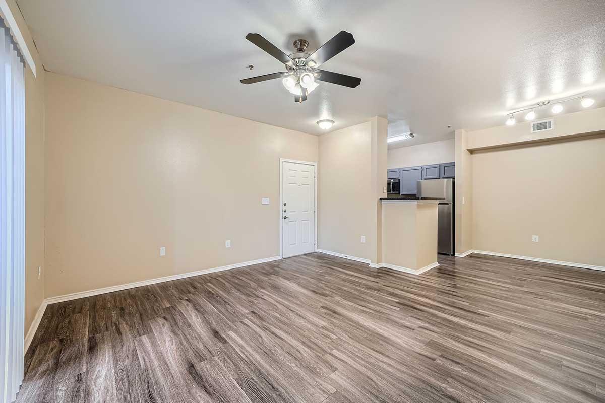 A spacious, empty living room with light beige walls and laminate flooring. There is a ceiling fan with five blades, a door leading outside, and a doorway leading to a kitchen area with dark cabinets in the background. The space is well-lit with overhead lights and has windows covered by blinds.