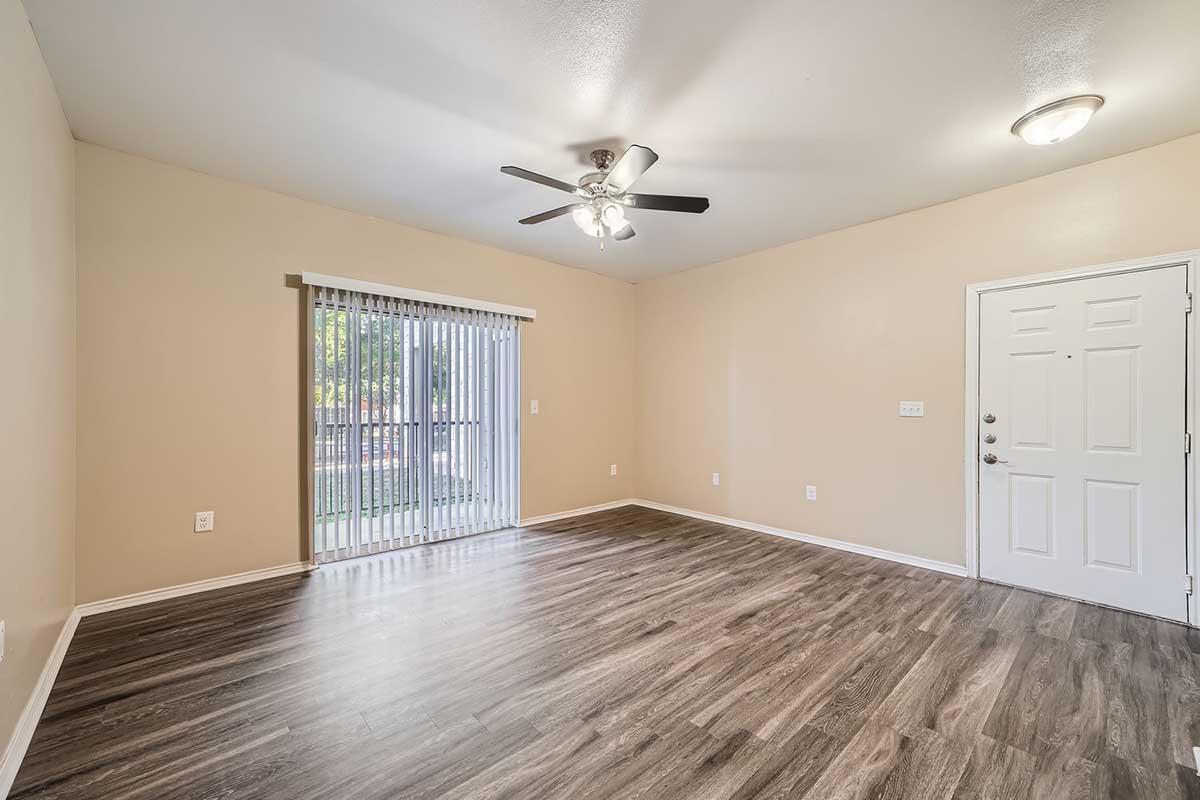 Empty living room with light-colored walls and hardwood-style flooring. A ceiling fan is mounted above, and a sliding glass door with vertical blinds leads to an outdoor area. The room has no furniture, providing a spacious and open feel, with a front door visible on the right.