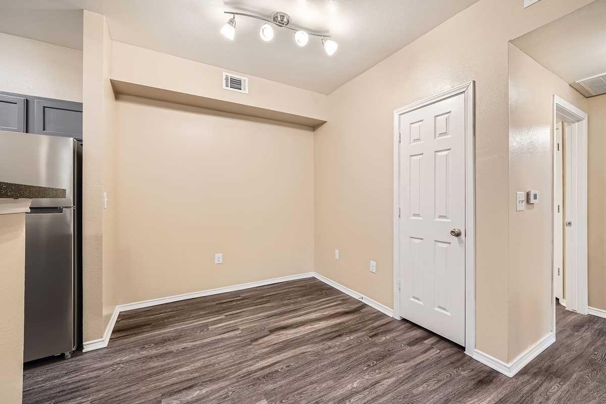 Interior of a small room with light beige walls and a dark wooden floor. There is a doorway on the right leading to another room and a closet with a white door. The ceiling has recessed lighting, and the kitchen area is visible on the left with dark cabinets. The space appears clean and empty, suitable for various uses.