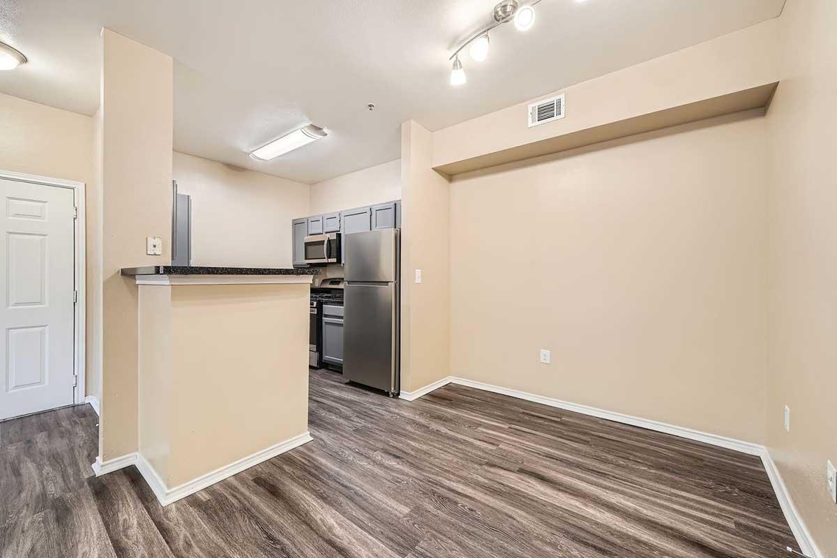 A small, modern kitchen area with an open floor plan. Features include light-colored walls, a dark countertop, grey cabinets, and stainless steel appliances. There’s a spacious living area with wood-style flooring and good lighting from overhead fixtures. A door is visible on the left, leading to another space.