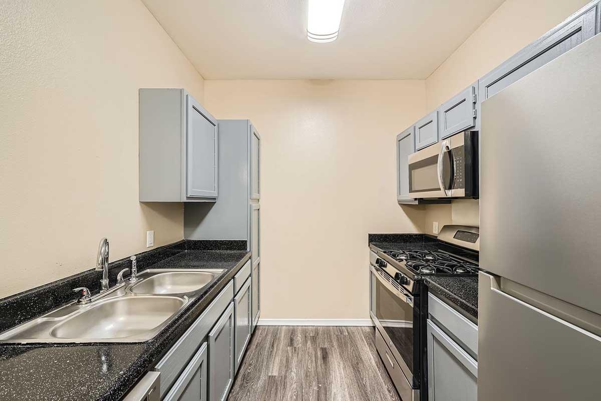 A modern kitchen featuring gray cabinets, a double sink, stainless steel appliances including a microwave and oven, and dark countertops. The walls are painted a light color, and the flooring is laminate, creating a clean and functional space.