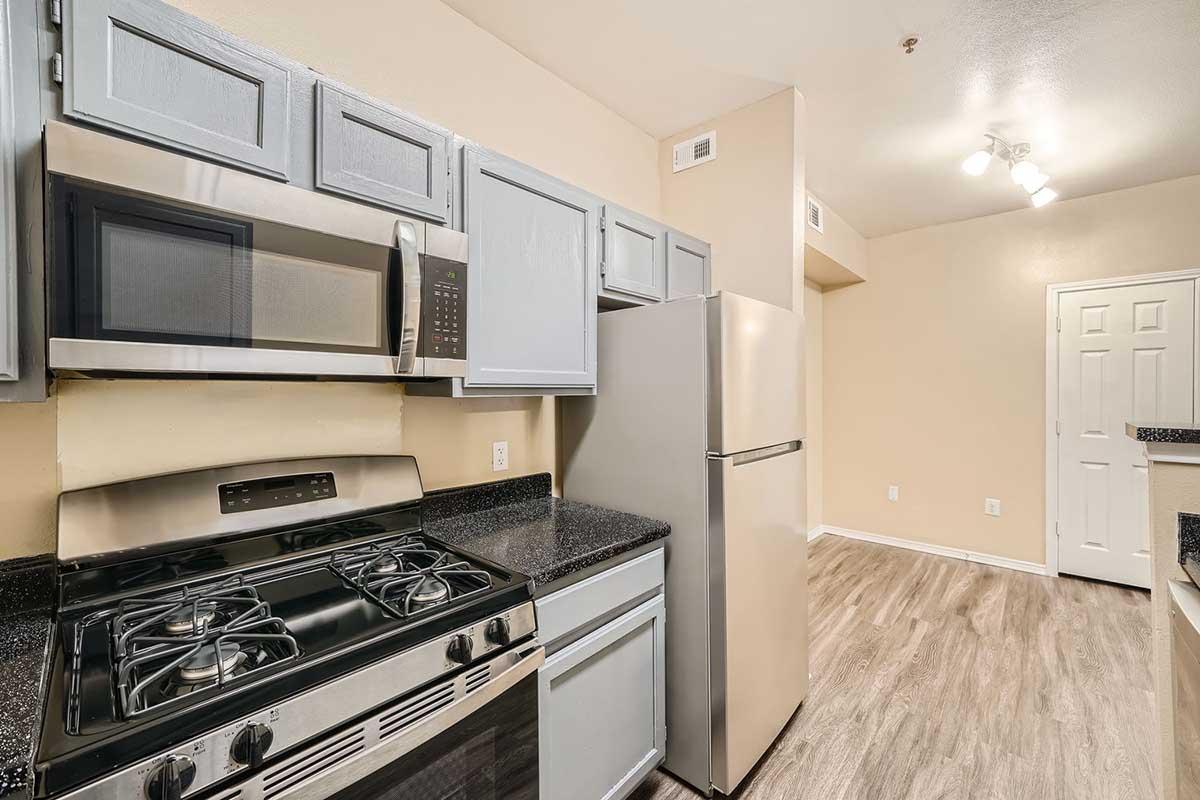Modern kitchen featuring light gray cabinets, a stainless steel refrigerator, and a gas stove. The countertops are dark granite, and the walls are painted a soft beige. There is a microwave built into the cabinetry, and the flooring is a light wood laminate. A door to a closet is visible in the background.