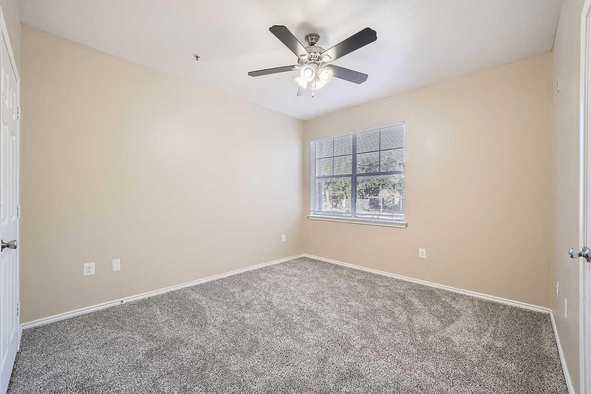 A vacant bedroom with light beige walls and gray carpet. The room features a ceiling fan with light fixtures, a window allowing natural light, and no furniture. The space appears clean and well-maintained, suitable for personal decoration and furnishing.