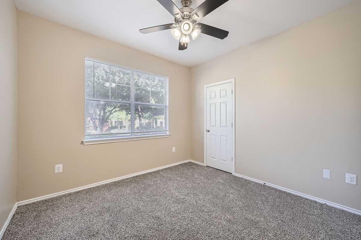 Empty room with light beige walls and a medium brown carpet. A ceiling fan with five blades is turned off. A window with blinds lets in natural light, and there's a white door on the right side of the image. The room appears clean and well-maintained.