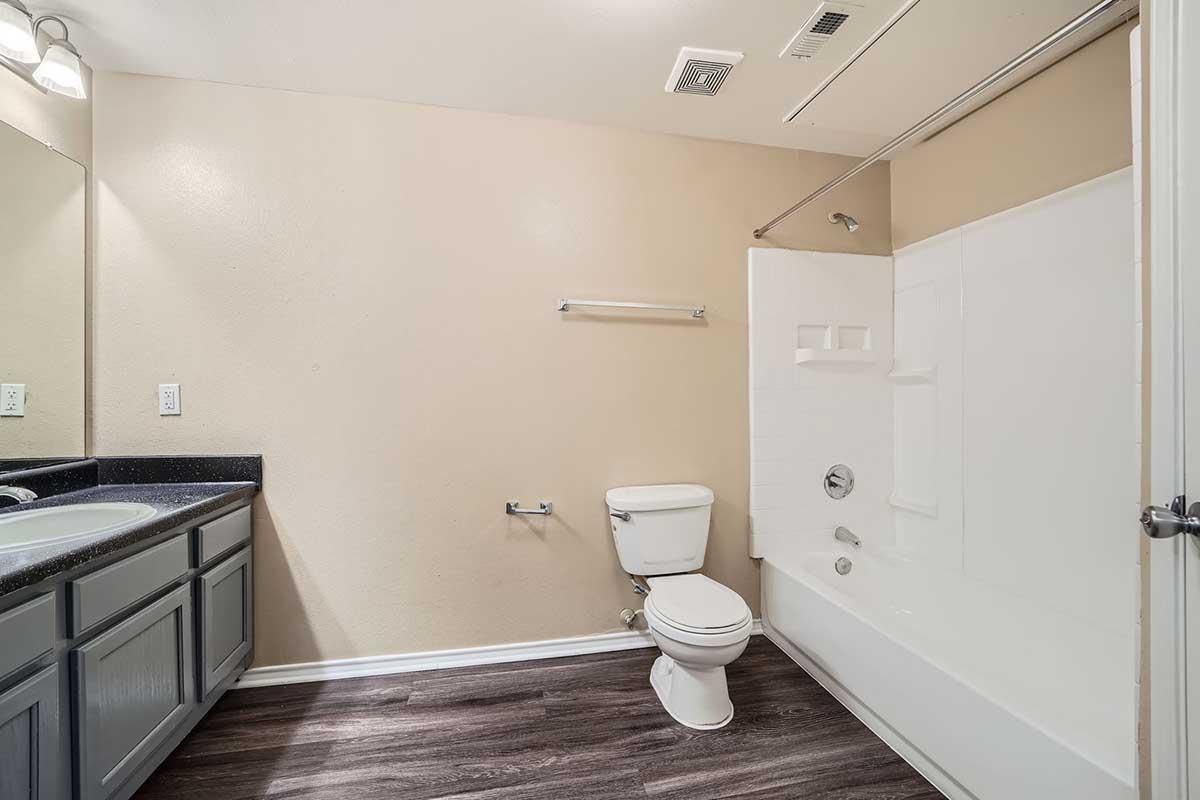 A clean, well-lit bathroom featuring a white bathtub with a shower, a toilet, and a dark countertop with two sinks. The walls are painted a light beige color, and there is a mirror above the sinks. The floor has dark wooden laminate flooring. A towel bar is mounted on the wall.