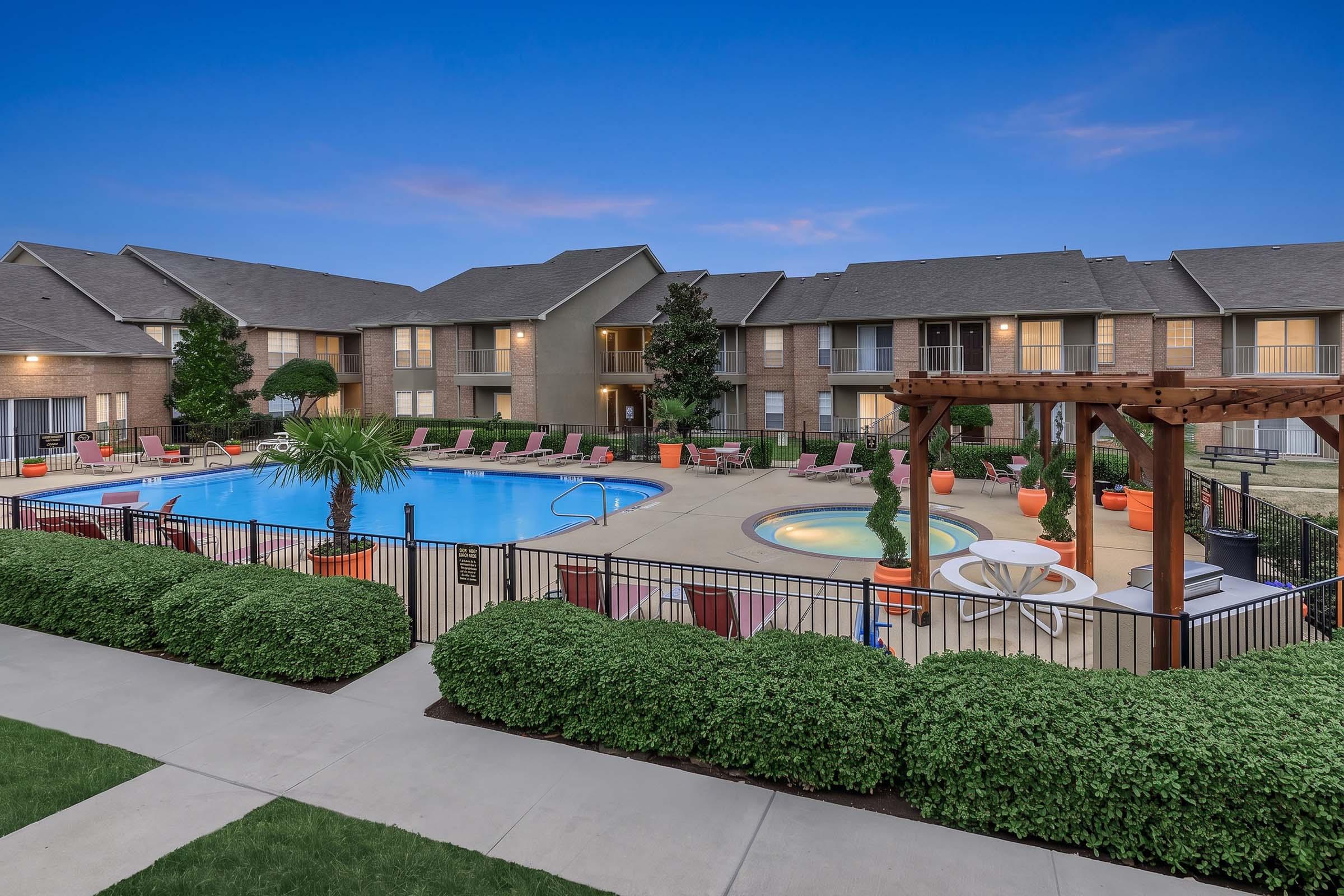A view of a well-maintained apartment complex showing a swimming pool surrounded by lounge chairs. There are landscaped green areas, including shrubs and trees. In the background, several apartment buildings are visible, and the sky is a soft blue with hints of twilight.