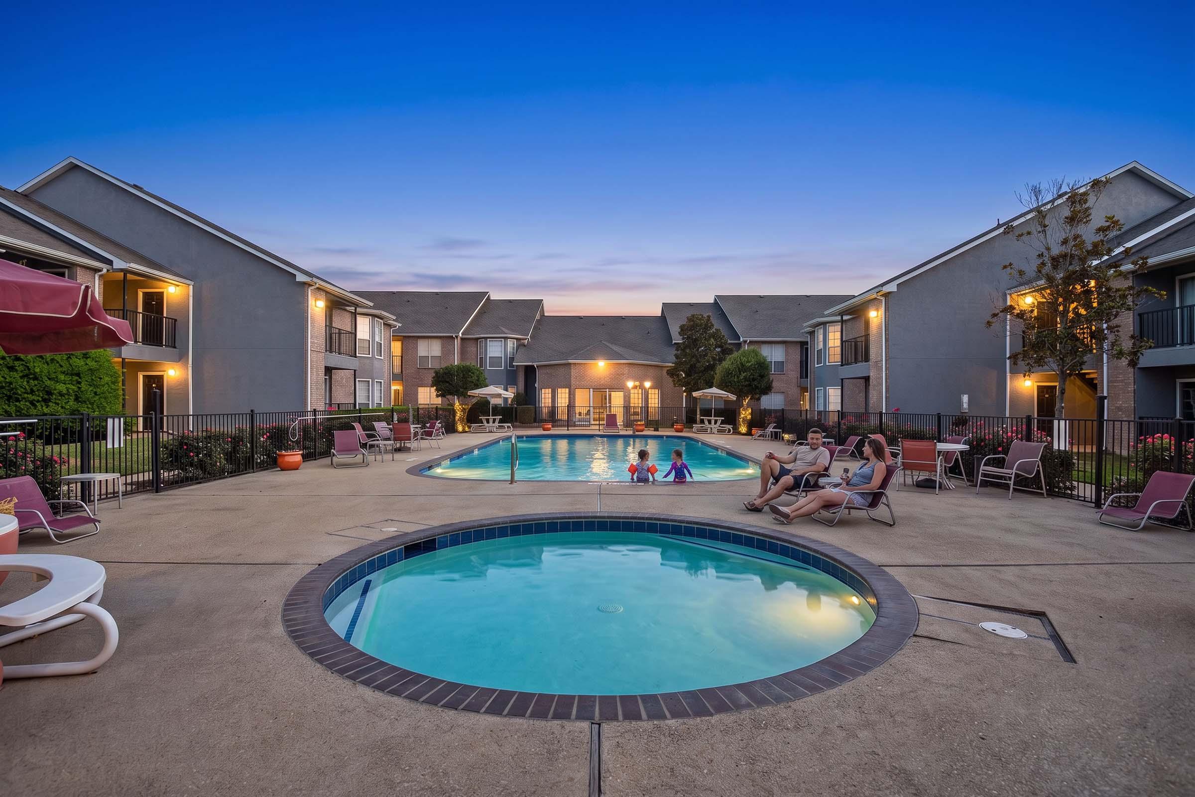 A tranquil outdoor swimming pool area surrounded by two-story apartment buildings. The main pool is large and inviting, with sun loungers nearby. A smaller hot tub is close to the pool. Two children are playing in the water while adults relax on chairs in the background, set against a beautiful evening sky.