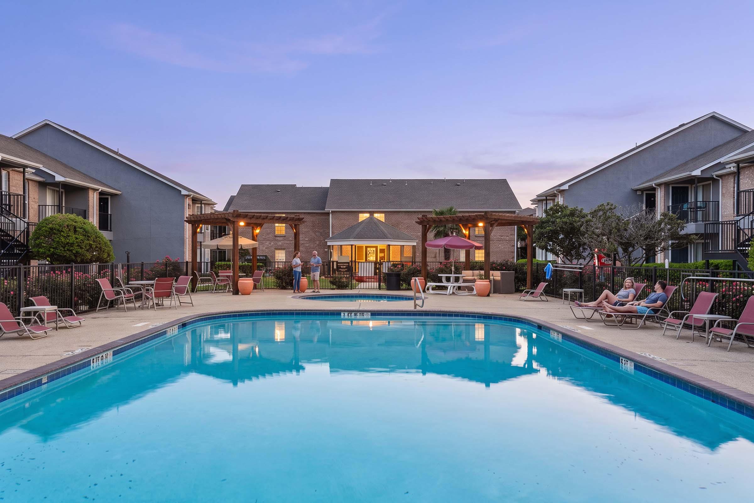 A serene view of a swimming pool surrounded by lounge chairs, with two apartment buildings in the background. The setting features vibrant greenery and a clear sky during dusk, reflecting calming blue tones in the water. People are enjoying the area, creating a relaxed atmosphere.