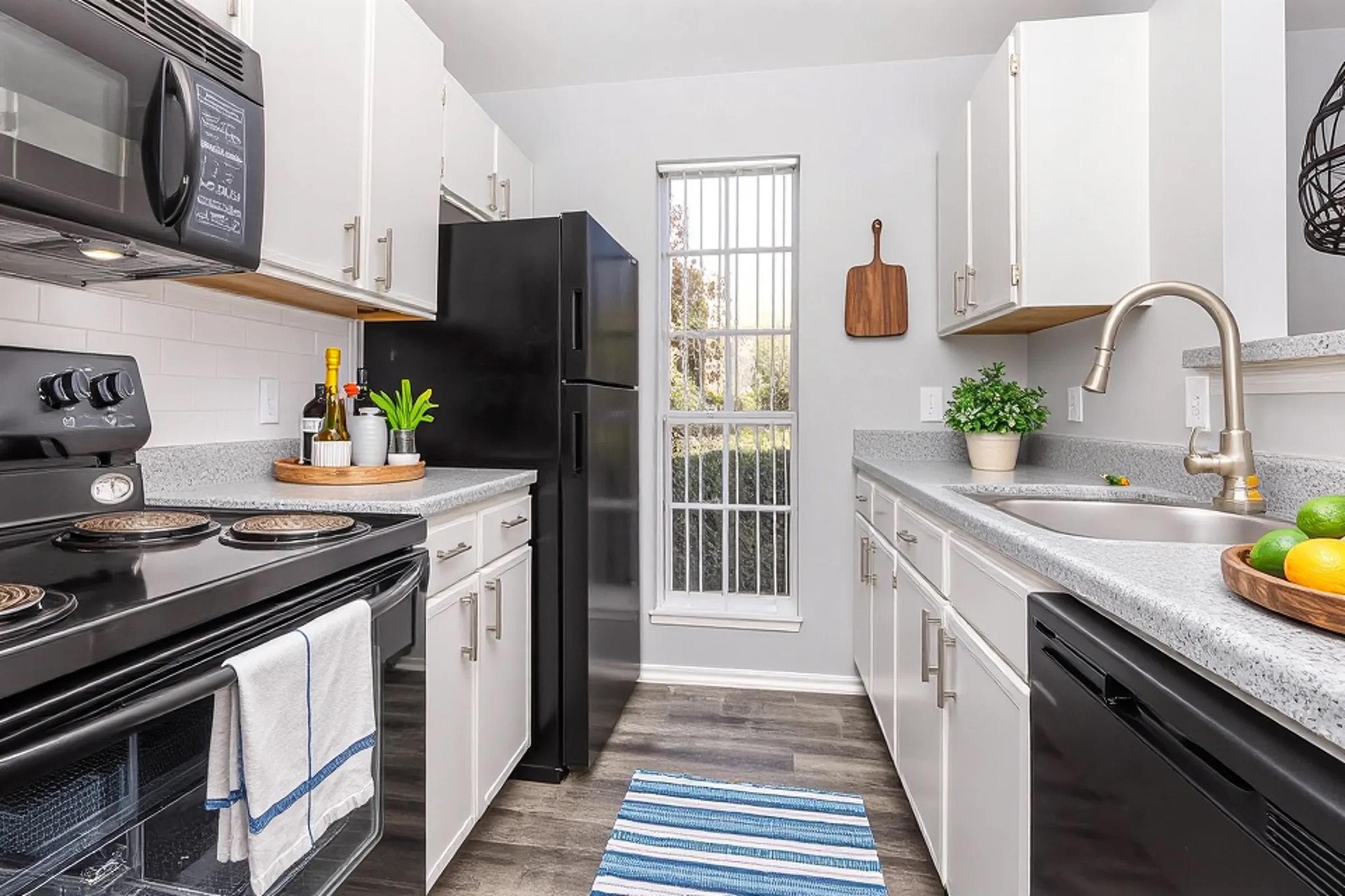 A modern kitchen featuring white cabinetry, a black refrigerator, and stainless steel appliances. The countertop is grey with a tray of green plants and fruits. A striped rug is on the floor, and a window provides natural light, enhancing the bright and airy atmosphere.