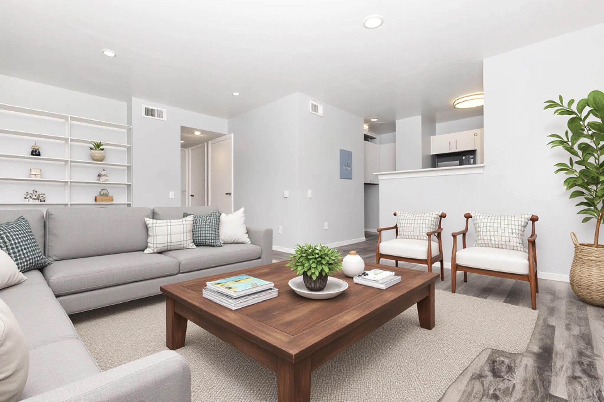 A modern living room with a light color scheme. It features a gray sectional sofa, two wooden accent chairs, a wooden coffee table with decor, and a potted plant. Bookshelves are on the left, and a glimpse of a kitchen can be seen in the background. The space is well-lit and inviting.