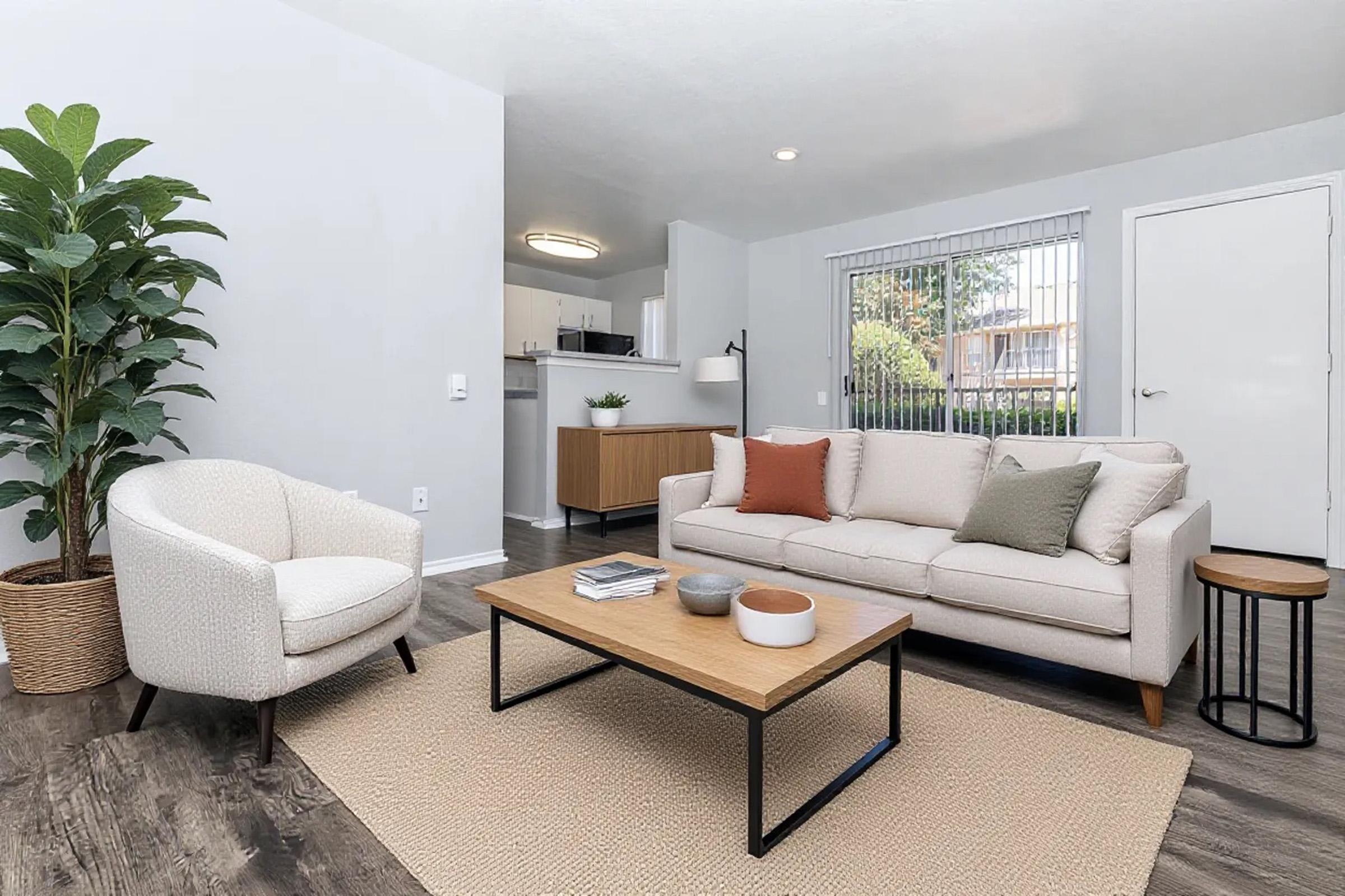 A cozy living room featuring a light-colored sofa with decorative pillows, a circular side table, and a textured area rug. A tall indoor plant adds greenery, and large windows provide natural light. The space features modern decor and a neutral color palette, creating a welcoming atmosphere.