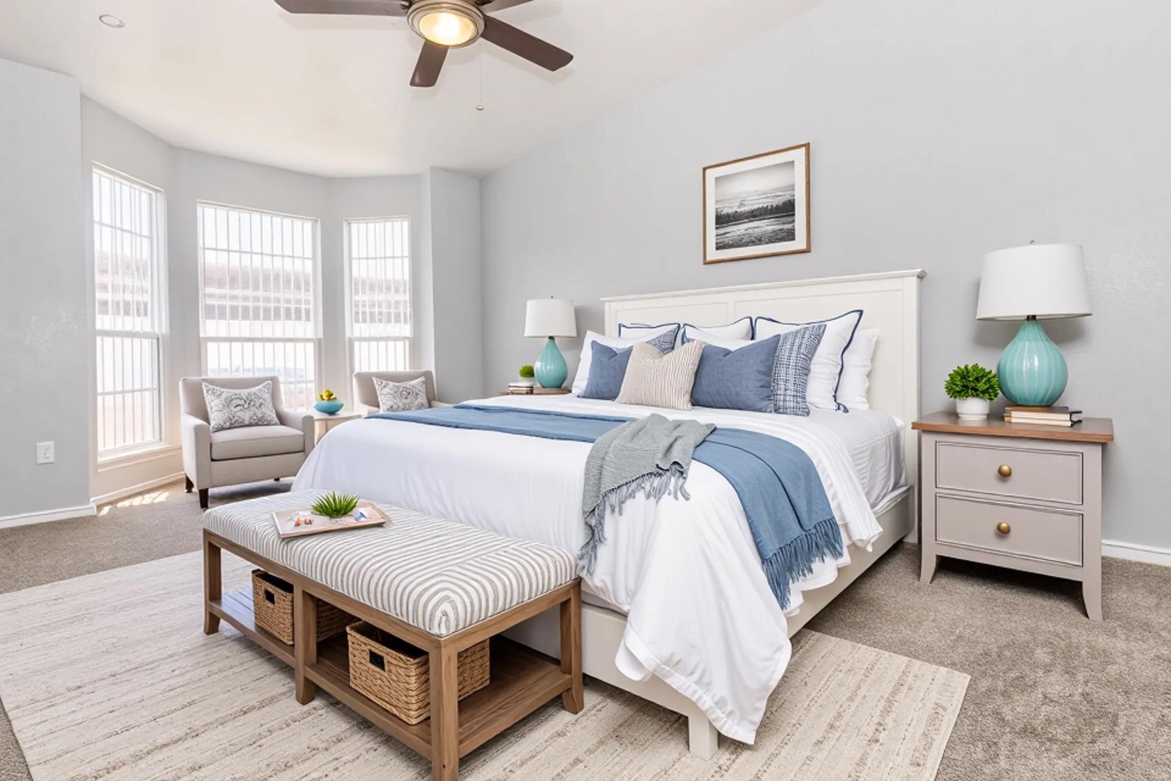 A cozy bedroom featuring a large bed with white and light blue bedding, two bedside tables with lamps, a bench with storage, and a comfortable seating area by the windows. The walls are painted light gray, and decorative elements include plants and framed wall art. Bright natural light fills the space.