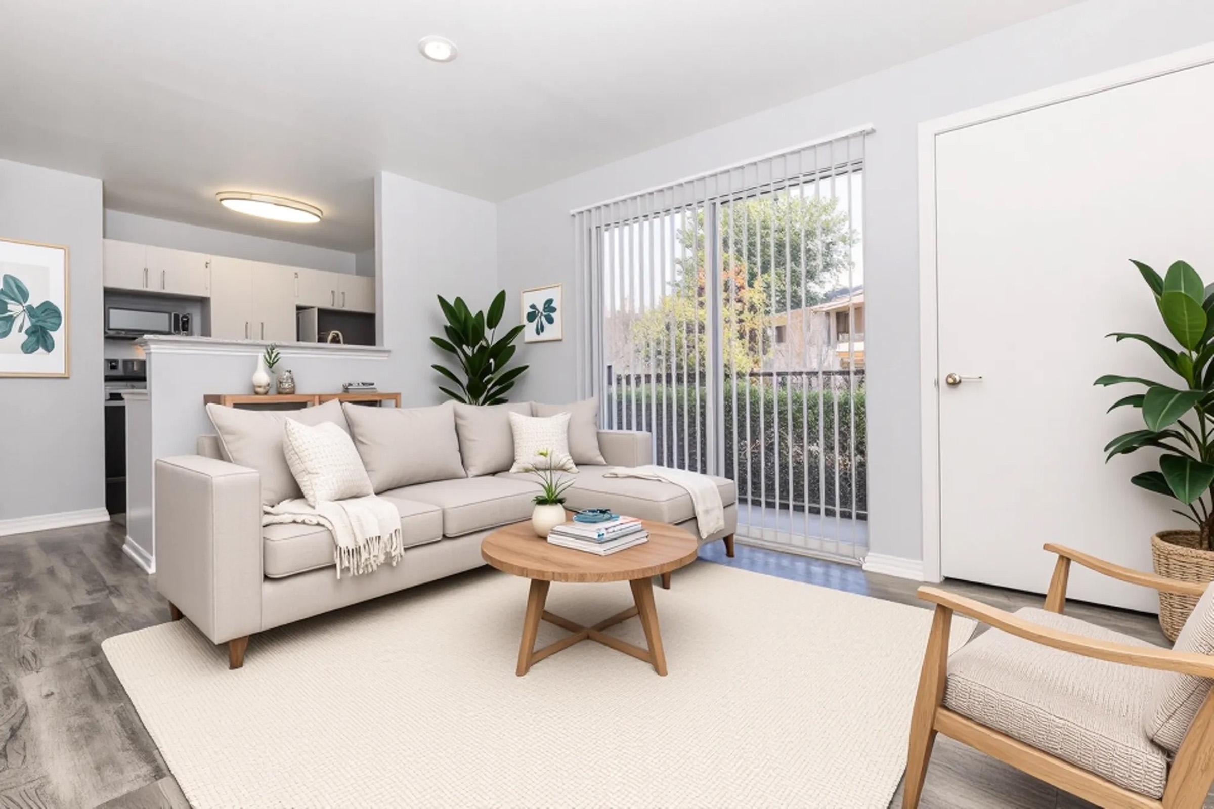A modern living room featuring a light gray sofa with cushions, a round wooden coffee table, and potted plants. There are framed leaf prints on the walls, a sliding door leading to the outdoors, and a beige area rug on a light wooden floor. A cozy armchair completes the space.