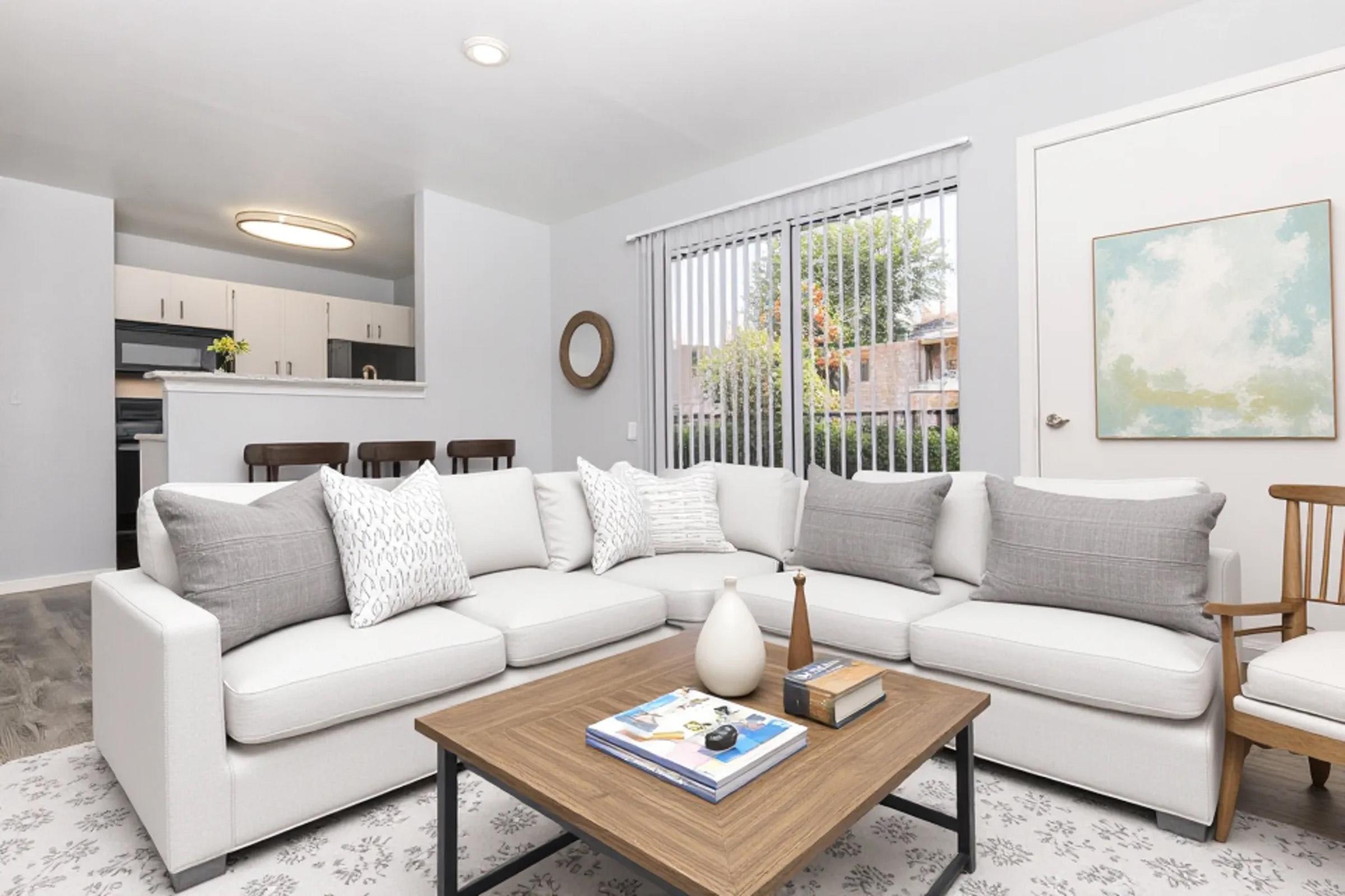 A modern living room featuring a large sectional sofa with gray and white cushions, a wooden coffee table with decorative items, and natural light coming through sliding glass doors. The space is decorated with a round mirror and a piece of abstract wall art, creating a cozy and inviting atmosphere.
