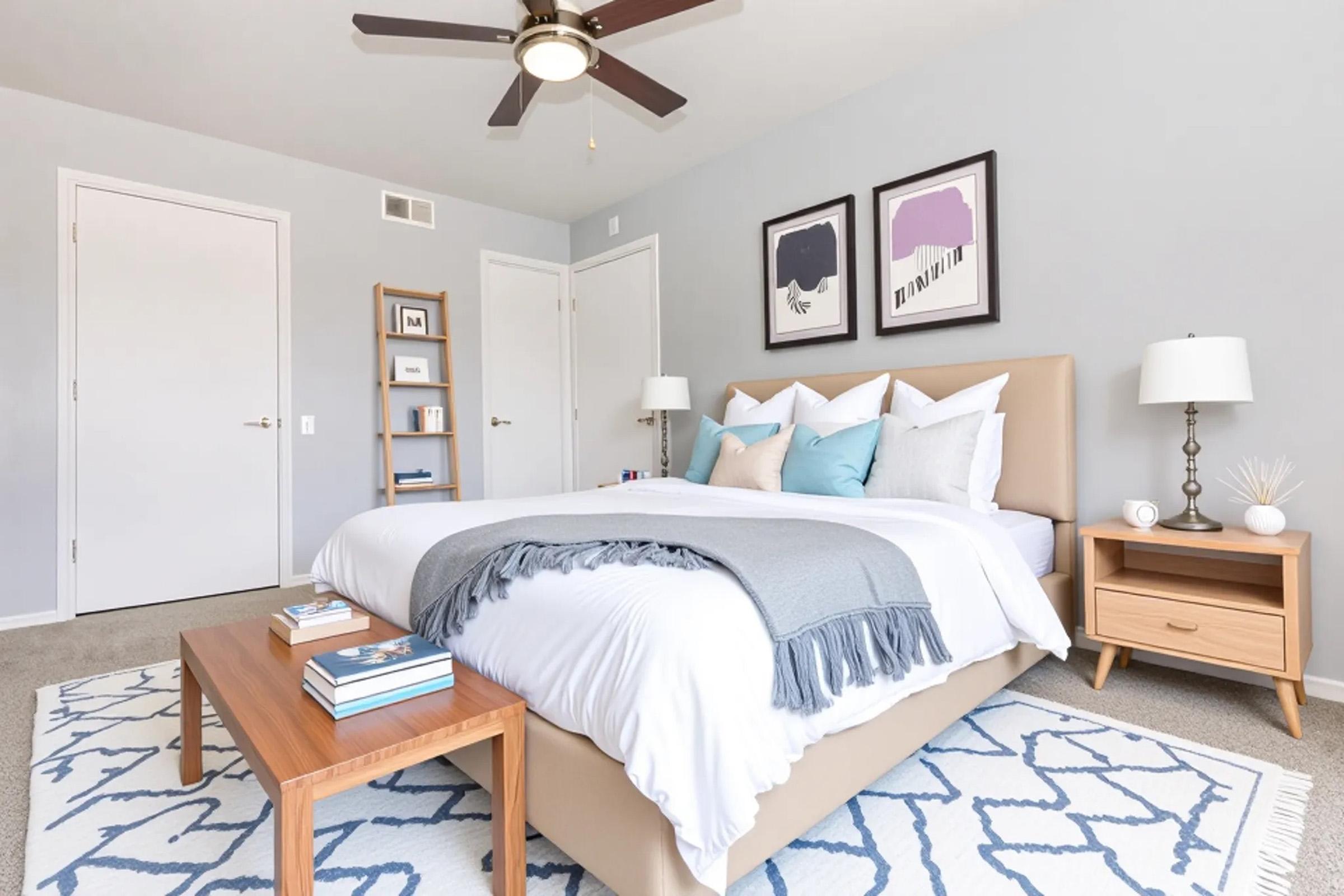 A modern, cozy bedroom featuring a large bed with white bedding and accent pillows in light blue. There are two framed artworks above the bed, a fan on the ceiling, and bedside tables with lamps. A wooden bookshelf and a patterned rug add a stylish touch to the room.