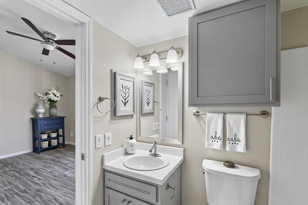 A modern bathroom featuring gray cabinetry, a stylish mirror with three light fixtures above, and a sink with soap dispenser. White towels hang on a rack, and a toilet is visible. In the background, a spacious room with a blue chest of drawers and floral decor can be seen.