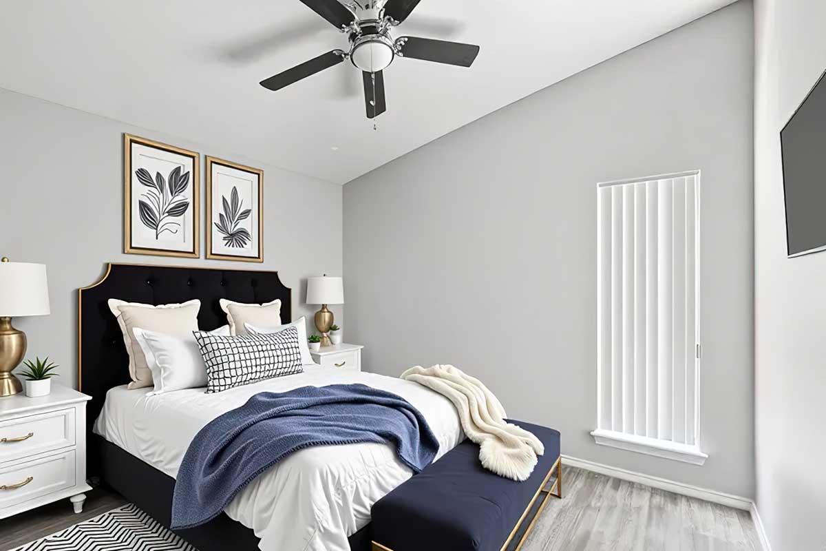 Cozy modern bedroom featuring a black upholstered bed with decorative pillows, a blue throw blanket, and a wooden bench at the foot. Two lamps on nightstands and framed botanical prints decorate the walls. A ceiling fan and a vertical window with blinds complete the serene ambiance.