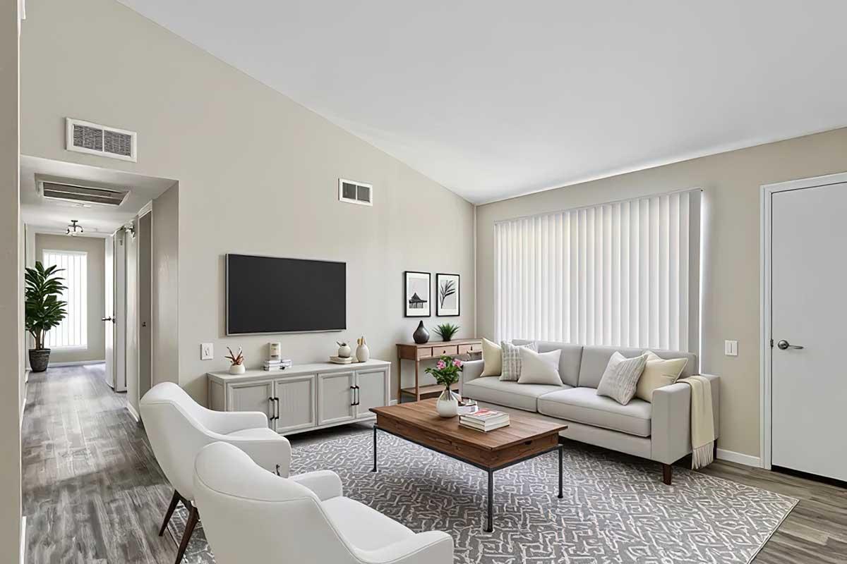 A modern, bright living room featuring a light gray sofa with decorative pillows, a wooden coffee table, two white armchairs, and a wall-mounted TV. The room has light-colored walls, a patterned area rug, and large windows with vertical blinds, creating an inviting and airy atmosphere.