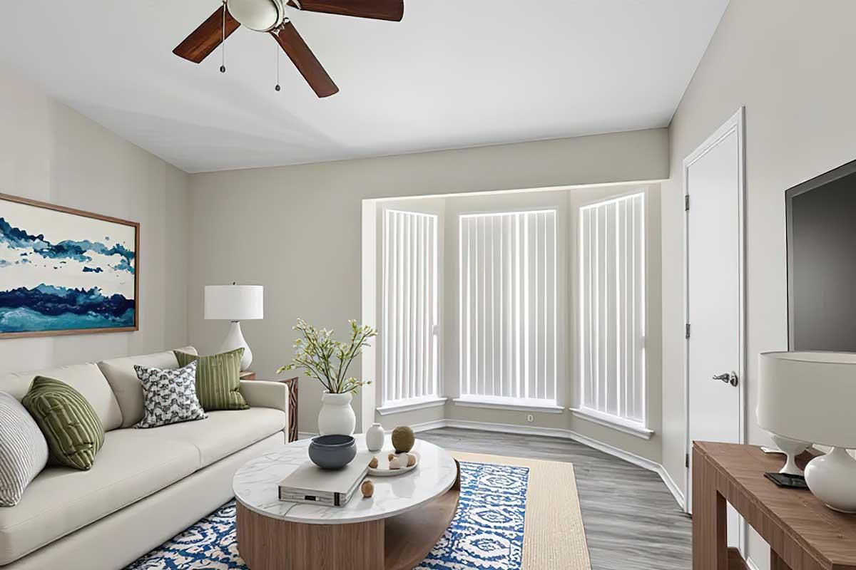 A modern living room featuring a light-colored sofa with decorative pillows, a round coffee table with assorted items, and a lamp. Large windows with vertical blinds allow natural light, and artwork is displayed on the wall. The flooring is a wood-like finish, complemented by a patterned area rug.