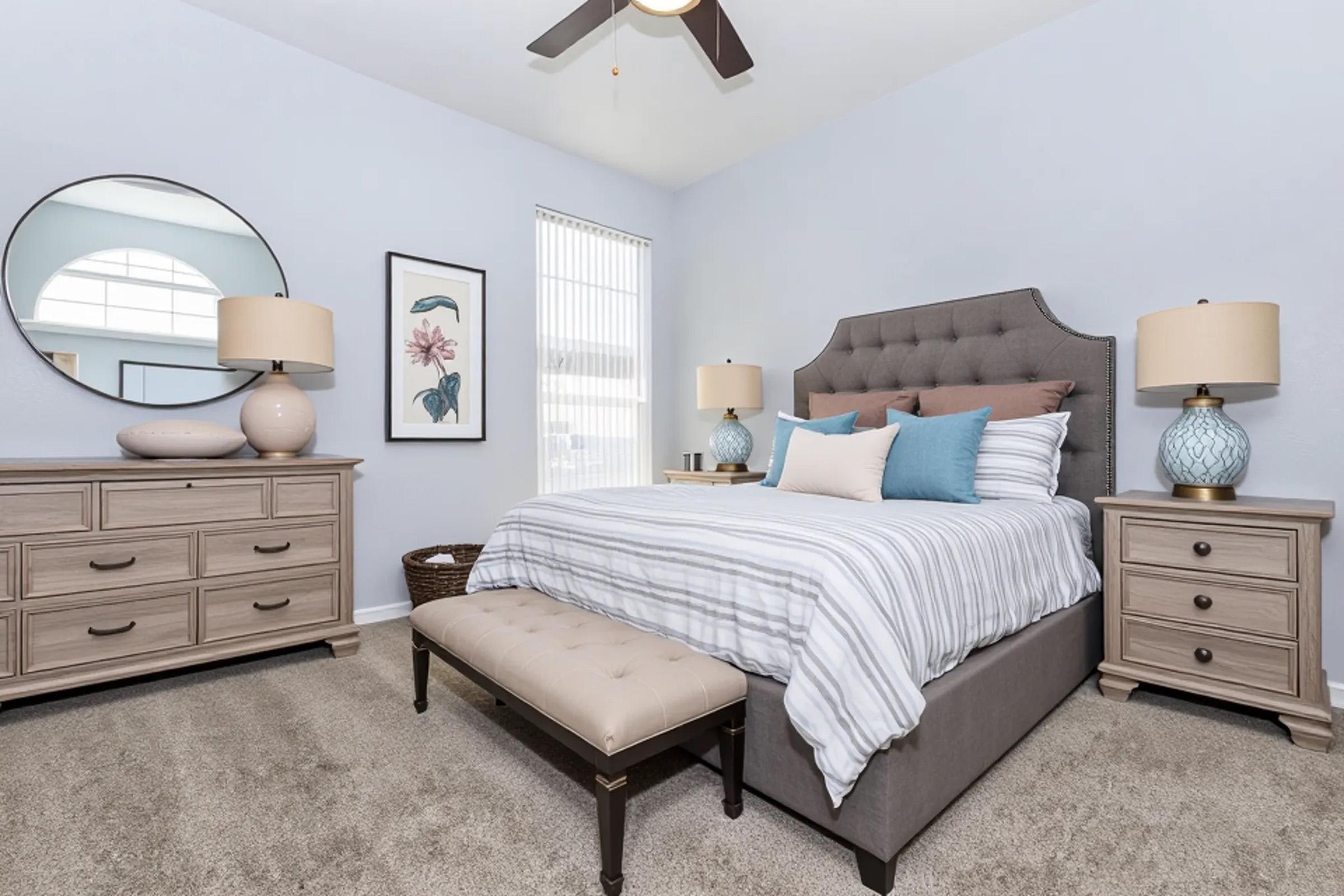 A cozy bedroom featuring a gray upholstered bed with striped bedding, blue accent pillows, and a padded bench at the foot. There are two stylish lamps on nightstands, a decorative mirror, and a dresser. Large windows allow natural light to brighten the space, complemented by soft, neutral colors and decorative artwork.