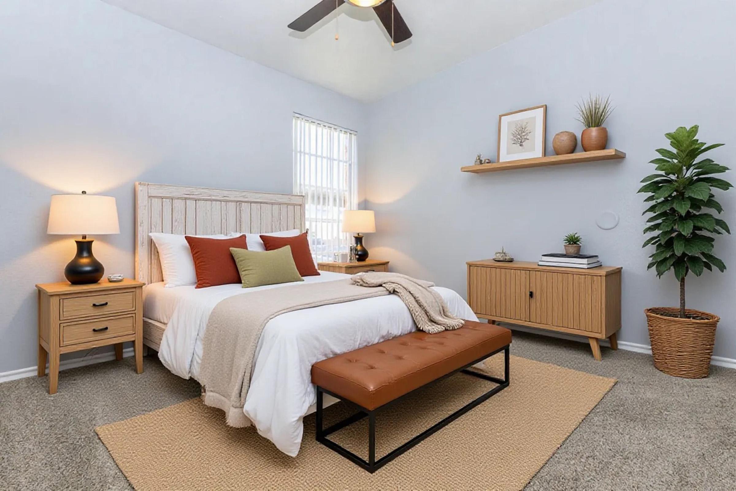 A cozy bedroom featuring a light-colored bed with decorative pillows, a bedside table with a lamp, and a wooden bench at the foot of the bed. A shelf with decorative items is mounted on the wall, and a potted plant adds a touch of greenery. The room has soft lighting and neutral tones.