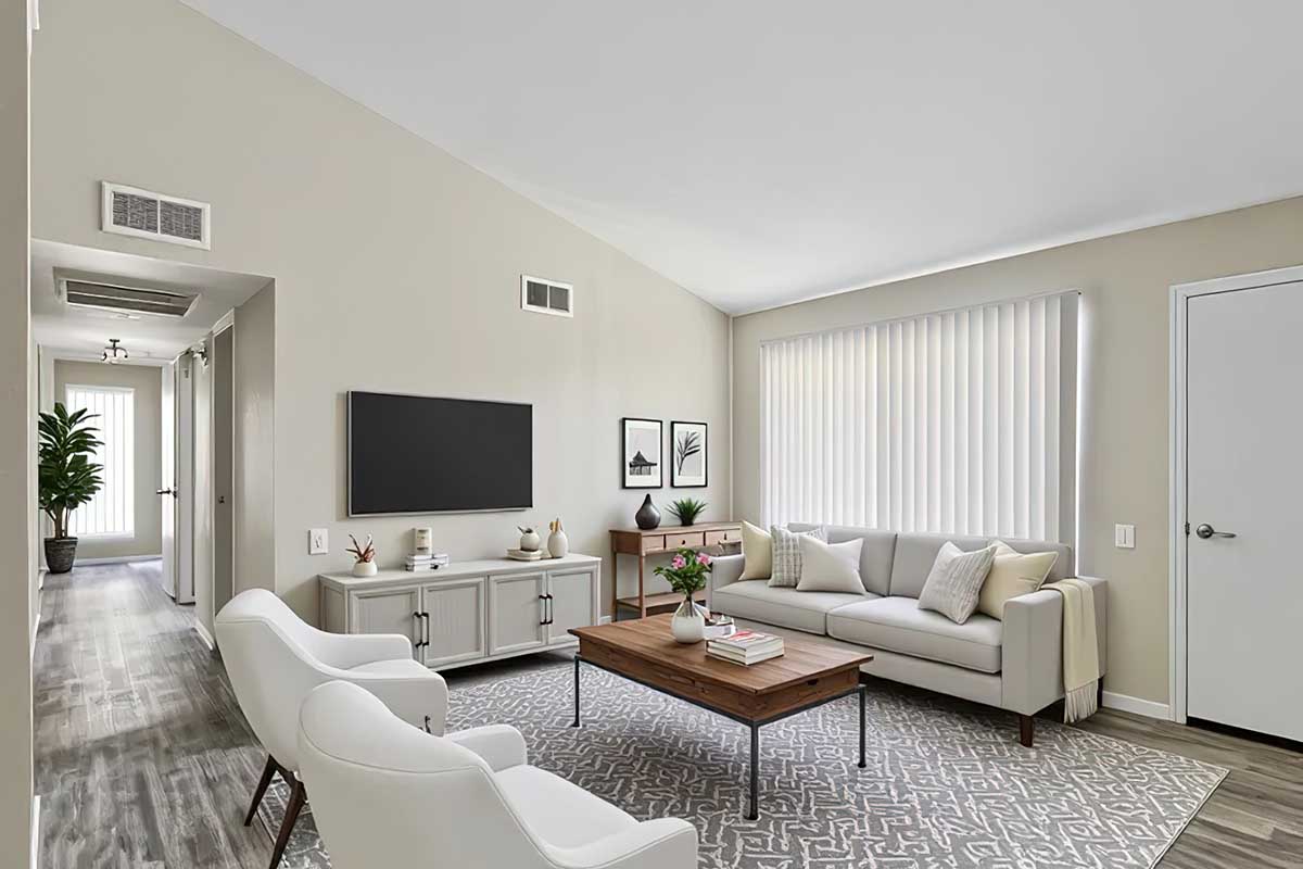A modern living room featuring a light gray sofa with decorative pillows, a wooden coffee table, and two white accent chairs. The space has neutral walls, a patterned rug, a wall-mounted TV, and a window with vertical blinds. A hallway with plants is visible in the background.
