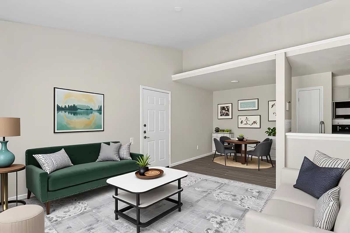 A bright and modern living room featuring a green couch, a coffee table, and decorative pillows. There's a lamp next to the couch, and wall art depicting a tranquil landscape. In the background, a dining area with a round table and chairs is visible, along with an open kitchen space. The walls are painted a soft neutral color.