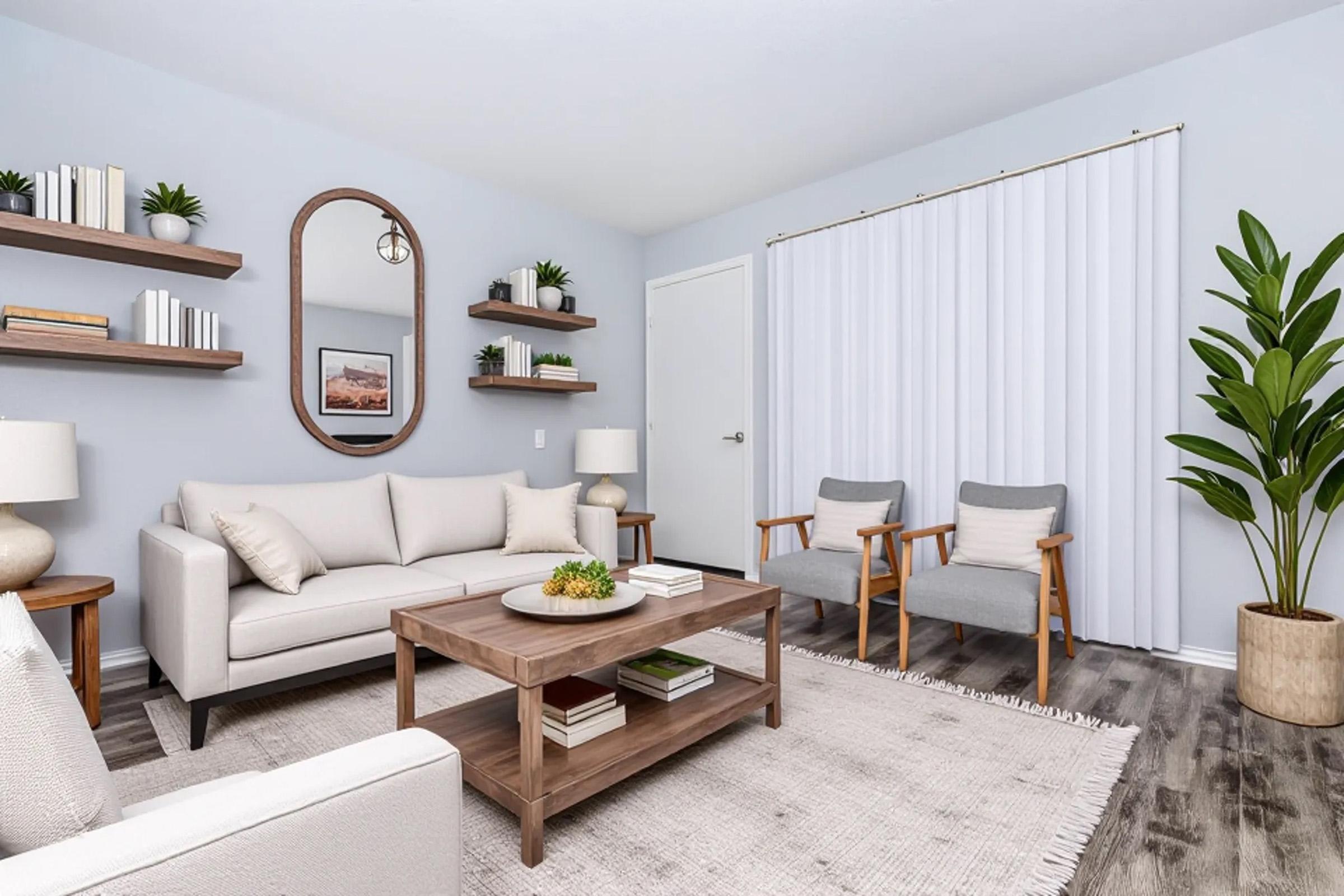 A modern living room featuring a light-colored sofa with decorative pillows, a wooden coffee table with a potted plant, and two gray armchairs. Wall-mounted shelves display books and decor, while a round mirror adds depth. Light-colored walls and a white vertical blind enhance the bright atmosphere.
