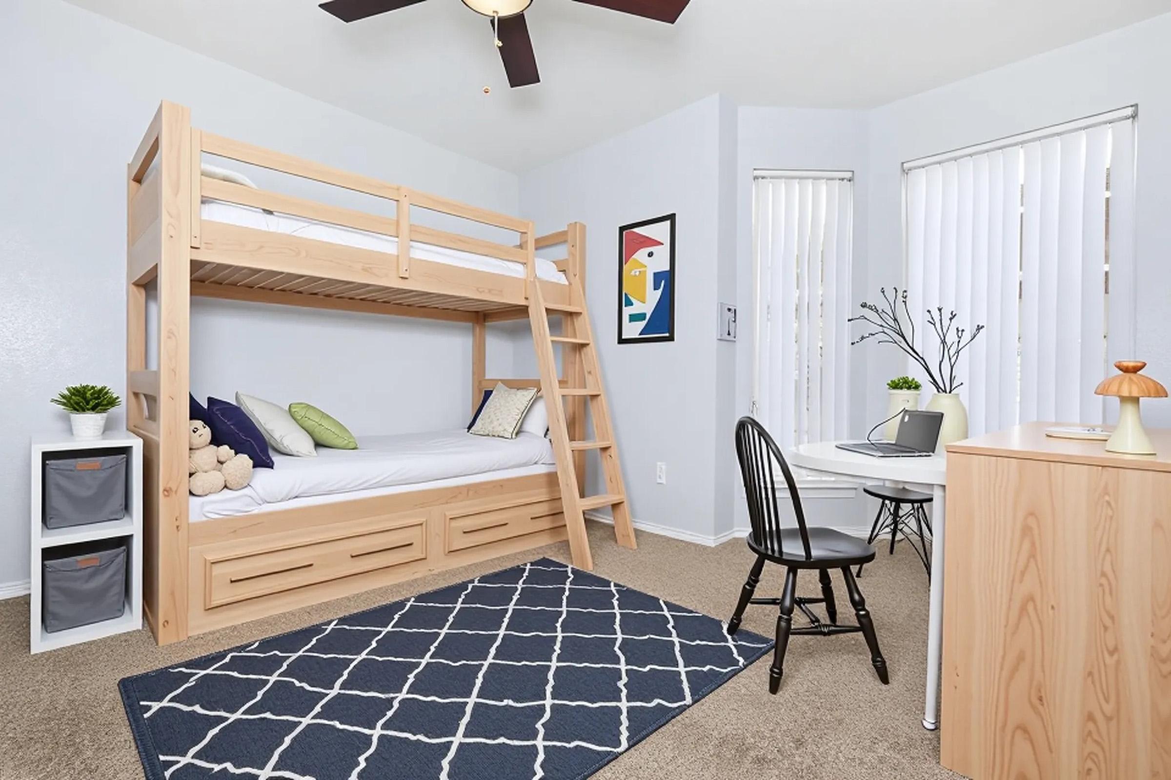 A cozy bedroom featuring a wooden bunk bed with a top and bottom sleeping area. A small desk with a chair is positioned nearby, along with a round table. The room has a modern decor style, with a blue geometric rug on the carpet and a few plants for added decor. Large windows provide natural light.