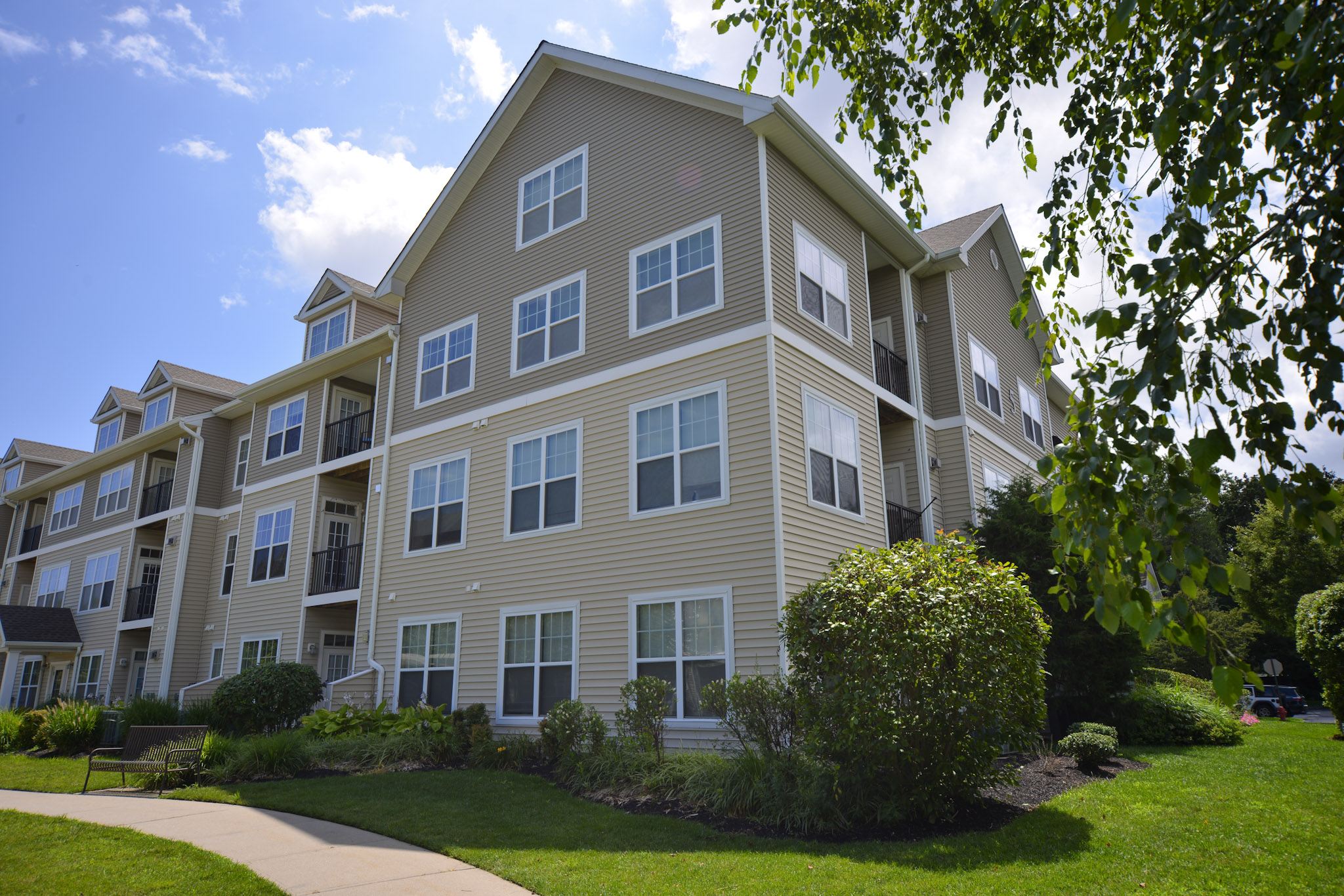 Modern multi-story apartment building with light tan siding, numerous large windows, and balconies. The surrounding landscape features well-maintained lawns, shrubs, and trees, under a bright blue sky with fluffy white clouds. A walking path leads alongside the building, enhancing the residential aesthetic.