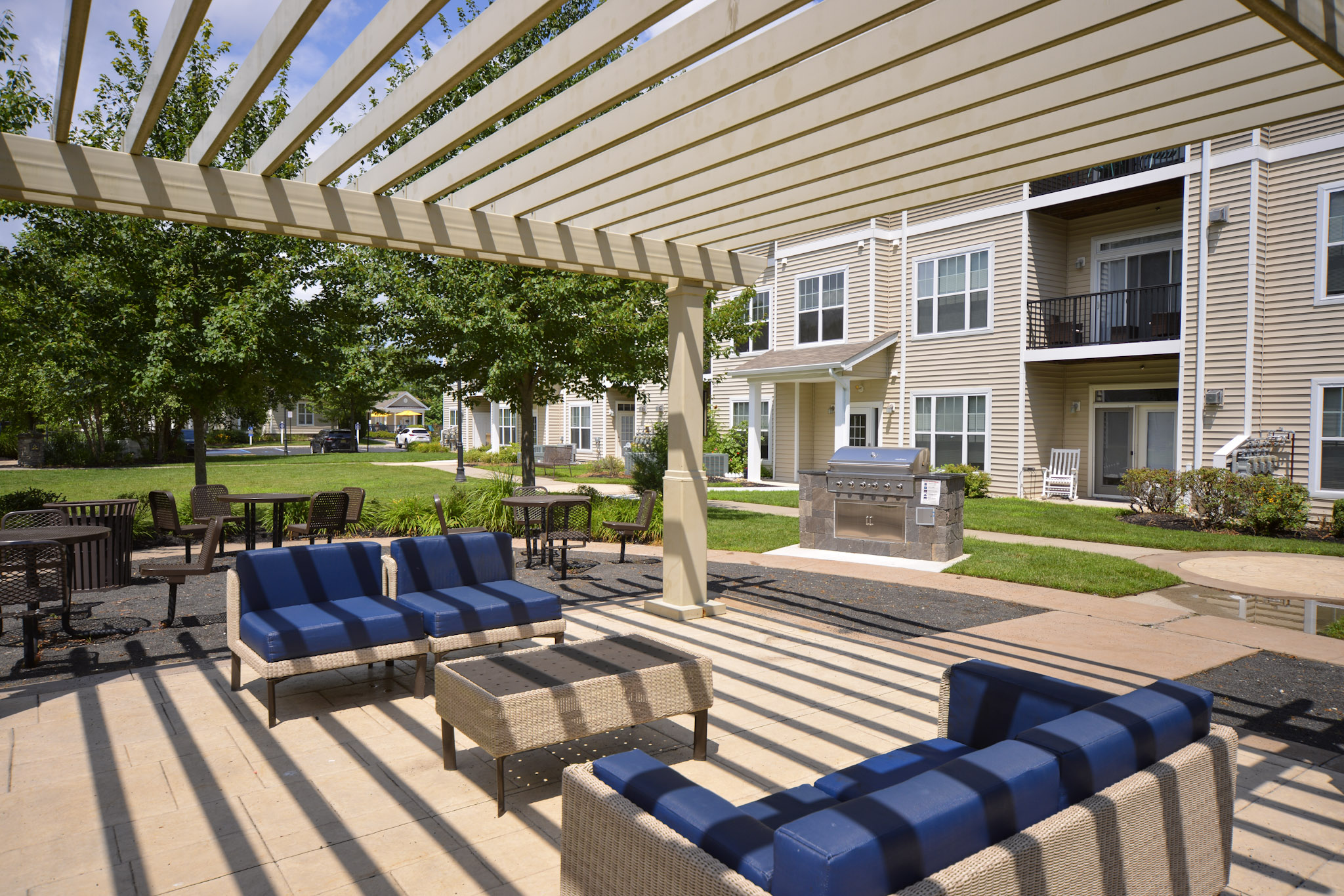 An outdoor communal area featuring modern seating arrangements with blue cushions under a slatted pergola, surrounded by green trees and well-maintained landscaping. In the background, there are apartment buildings and a barbecue grill nearby, creating a welcoming environment for relaxation.