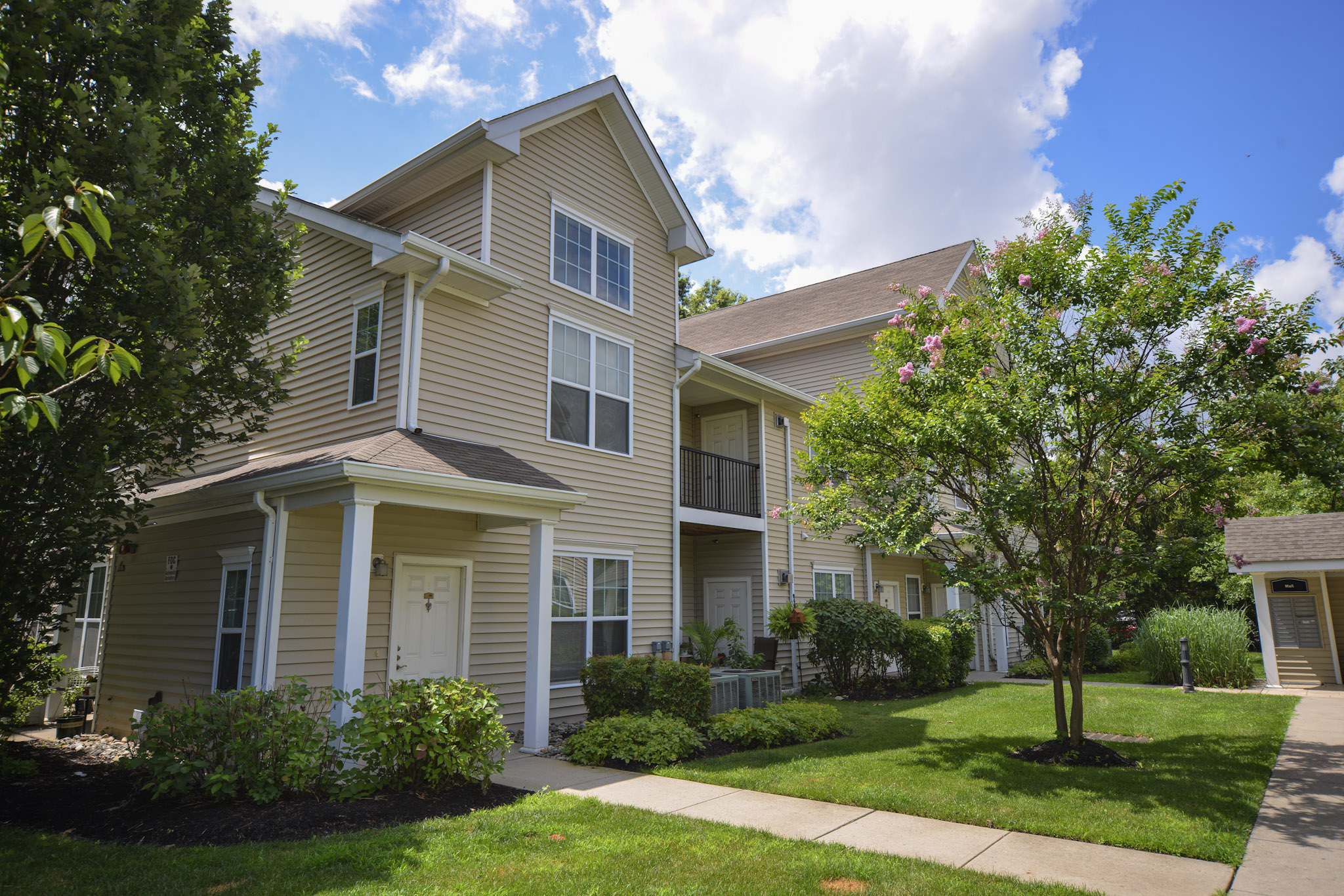 A two-story residential building with light beige siding, large windows, and balconies. The property features well-maintained landscaping, including bushes and trees. A paved walkway leads to the entrance, and the sky is partly cloudy, adding to the pleasant suburban atmosphere.