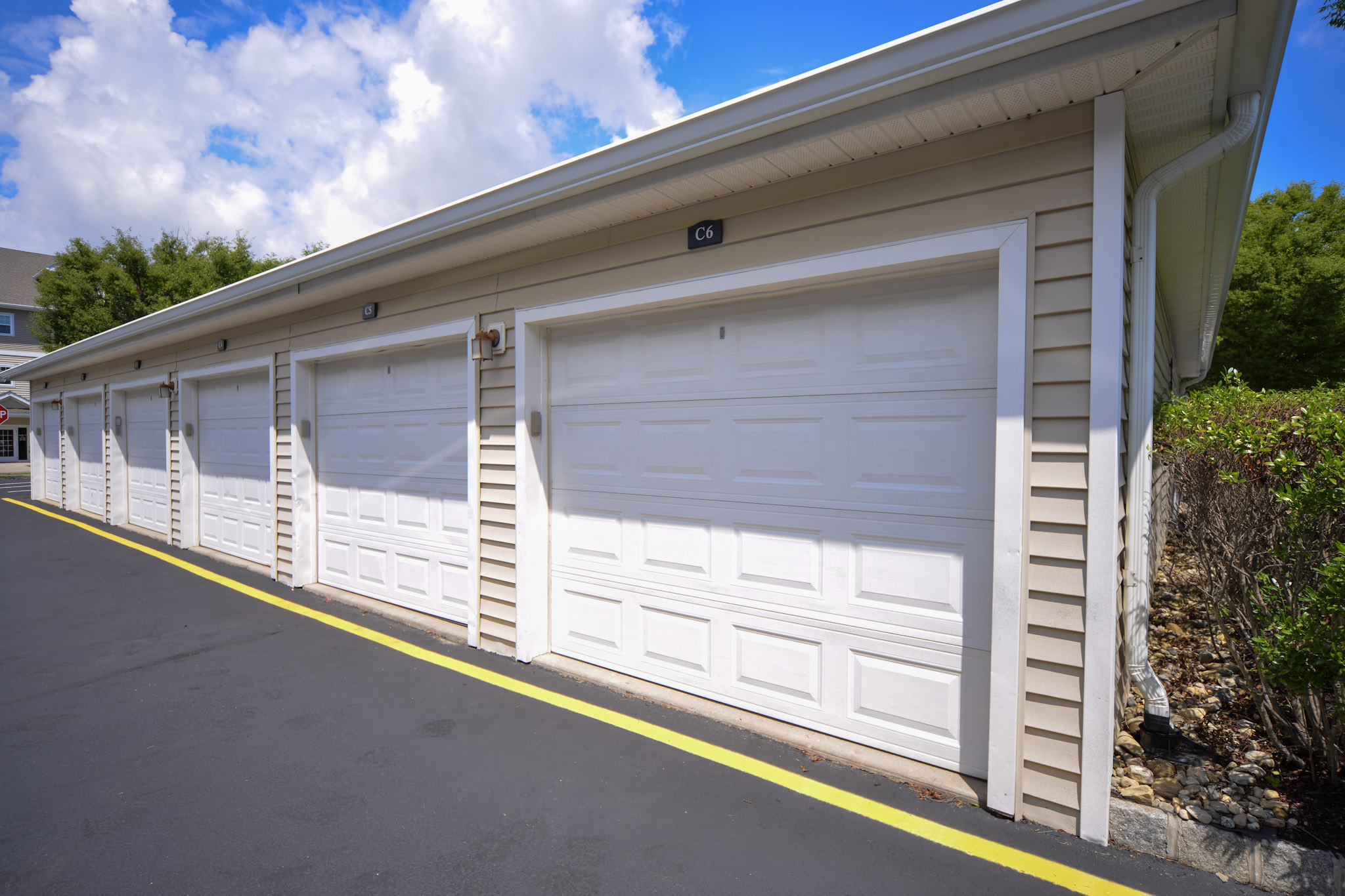 Row of closed white garage doors attached to a light-colored building. The garages are aligned neatly along a paved driveway, with a bright blue sky and fluffy clouds in the background. Yellow lines mark the edge of the driveway, adding contrast to the scene.