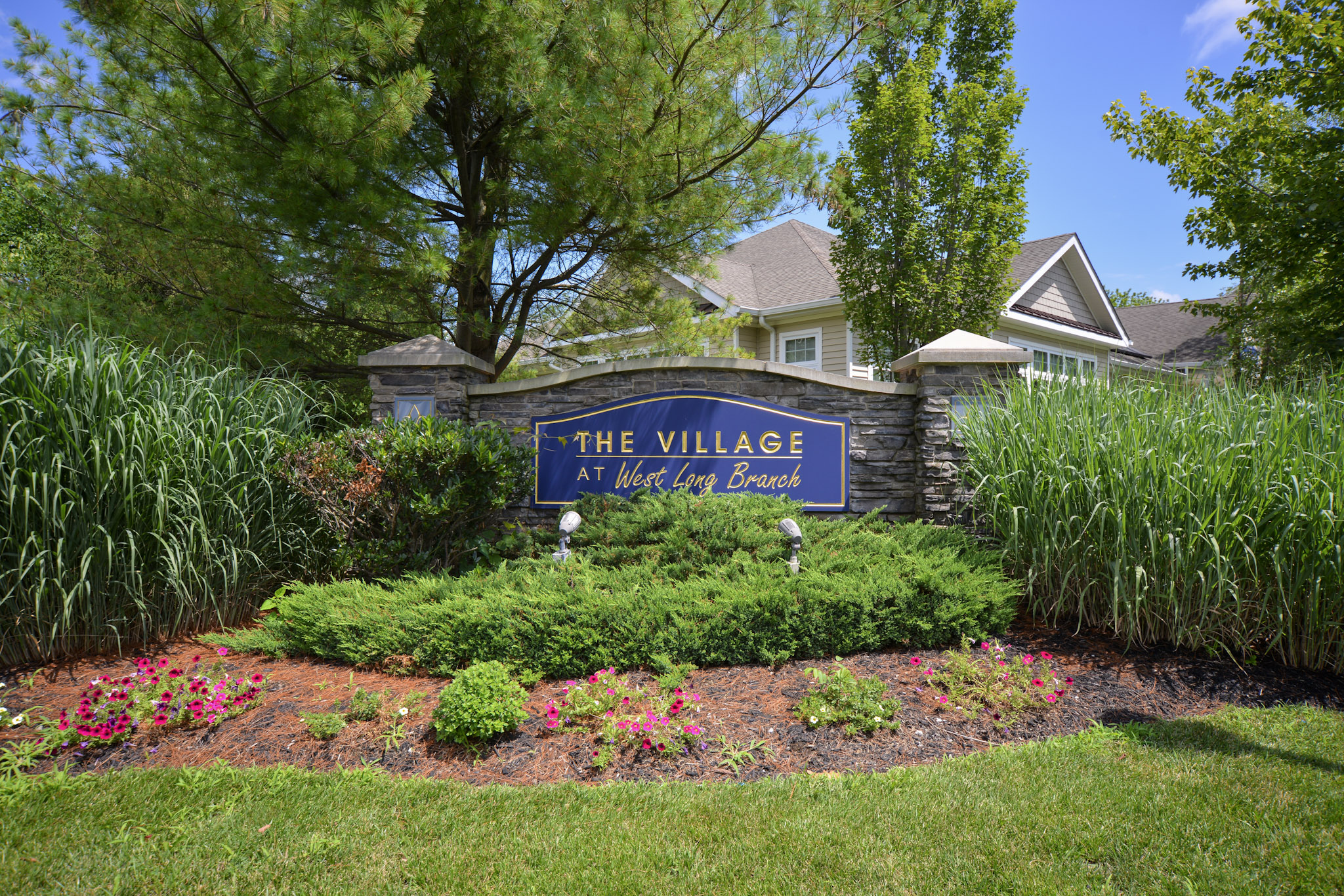 Sign for "The Village at West Lake Branch" surrounded by lush green grass, shrubs, and colorful flowers. A clear blue sky and trees are visible in the background, enhancing the welcoming atmosphere of the location. The sign features bold lettering and sits prominently at the entrance of the community.