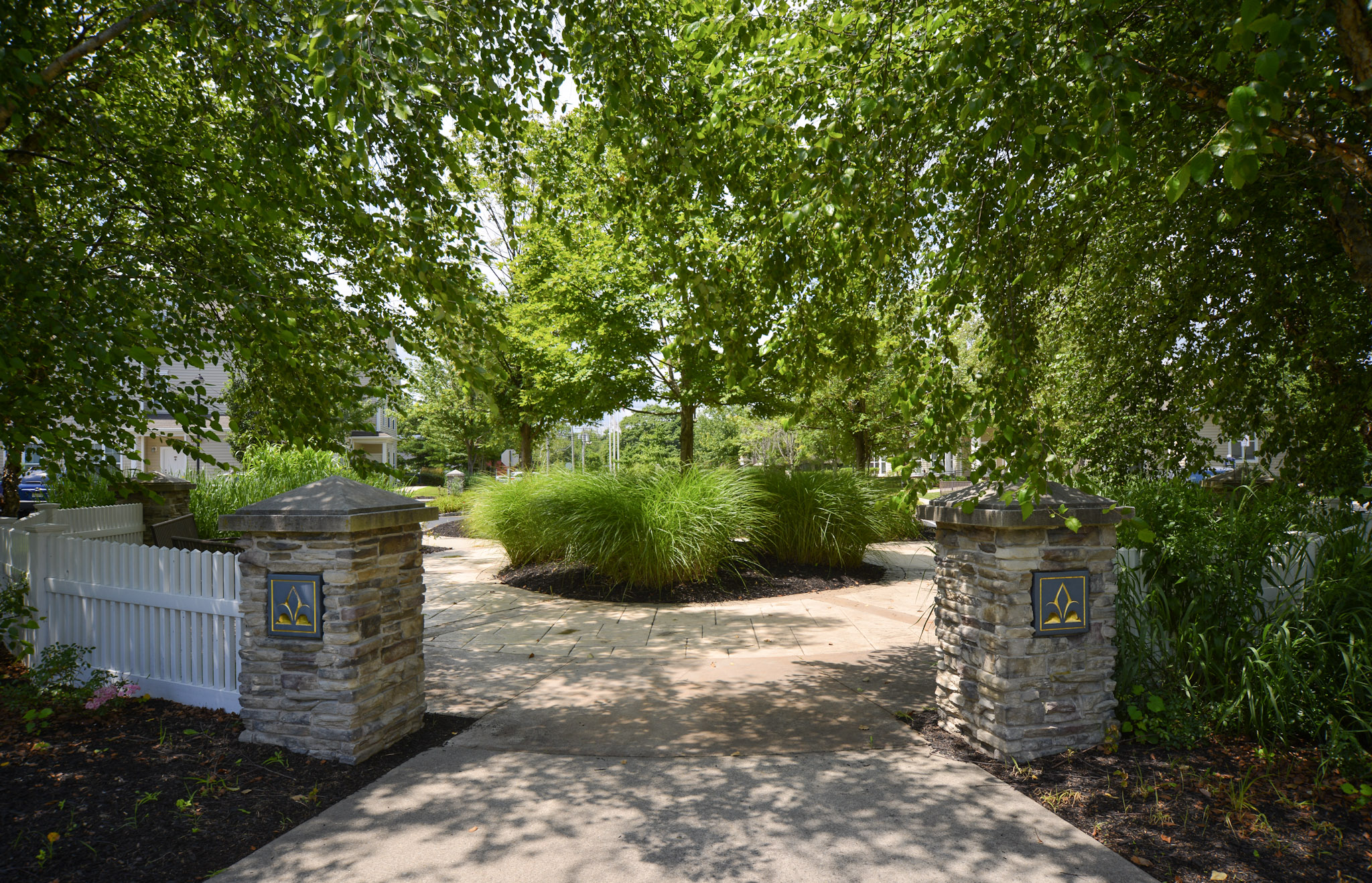 A tranquil garden pathway flanked by stone columns adorned with decorative emblems. Lush greenery, including tall grasses and trees, creates a peaceful atmosphere. A white picket fence surrounds the garden, enhancing the serene outdoor setting. The sunlight filters through the leaves, adding a warm glow to the scene.