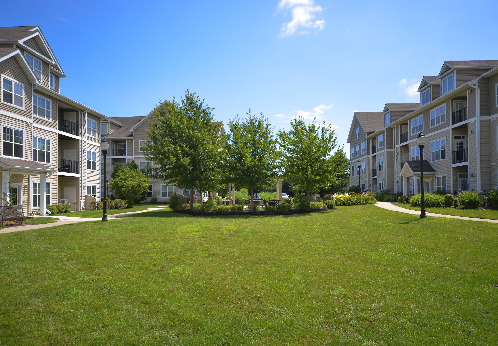 A sunny courtyard surrounded by residential apartment buildings. The grassy area features neatly trimmed lawns and small trees, with pathways leading through the space. Bright blue skies and a few fluffy clouds create a pleasant outdoor atmosphere.