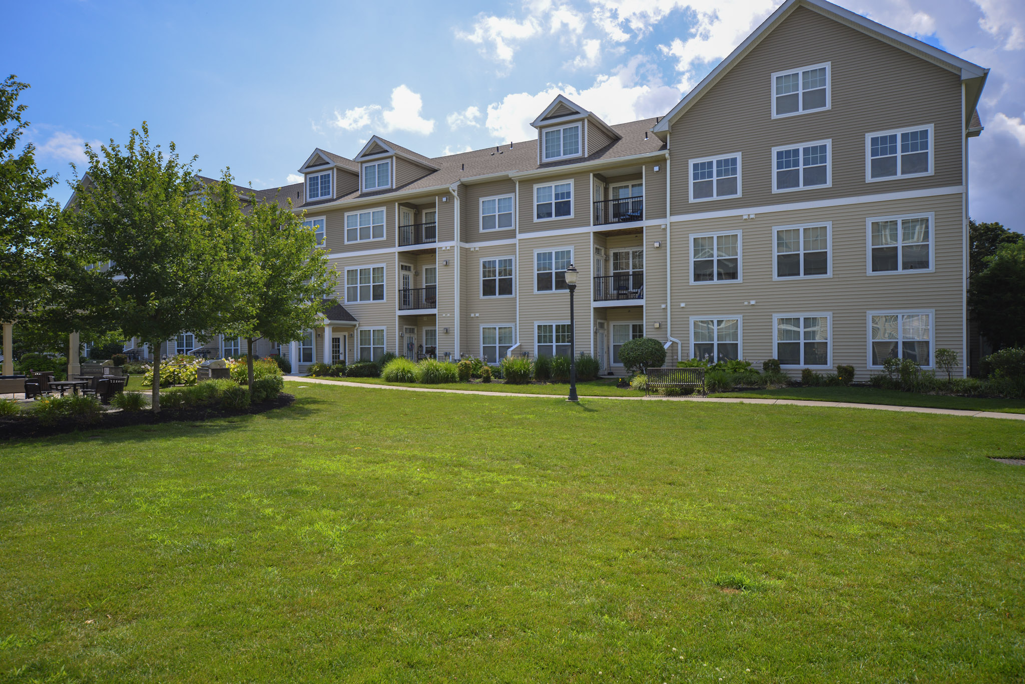 A well-maintained apartment building with multiple balconies, surrounded by green grass and landscaping. There are trees and shrubs in the foreground, and the sky is partly cloudy with some sunlight illuminating the scene.