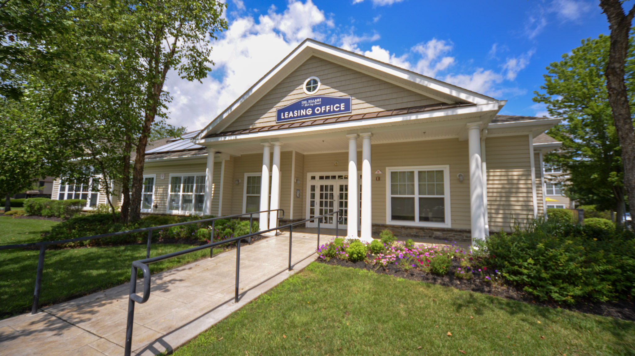 A leasing office building with a welcoming entrance, featuring a sign that reads "Leasing Office." The structure has a light-colored façade and a sloped roof, surrounded by greenery and flowers. A paved walkway with a railing leads up to the entrance, set against a backdrop of blue skies and fluffy clouds.