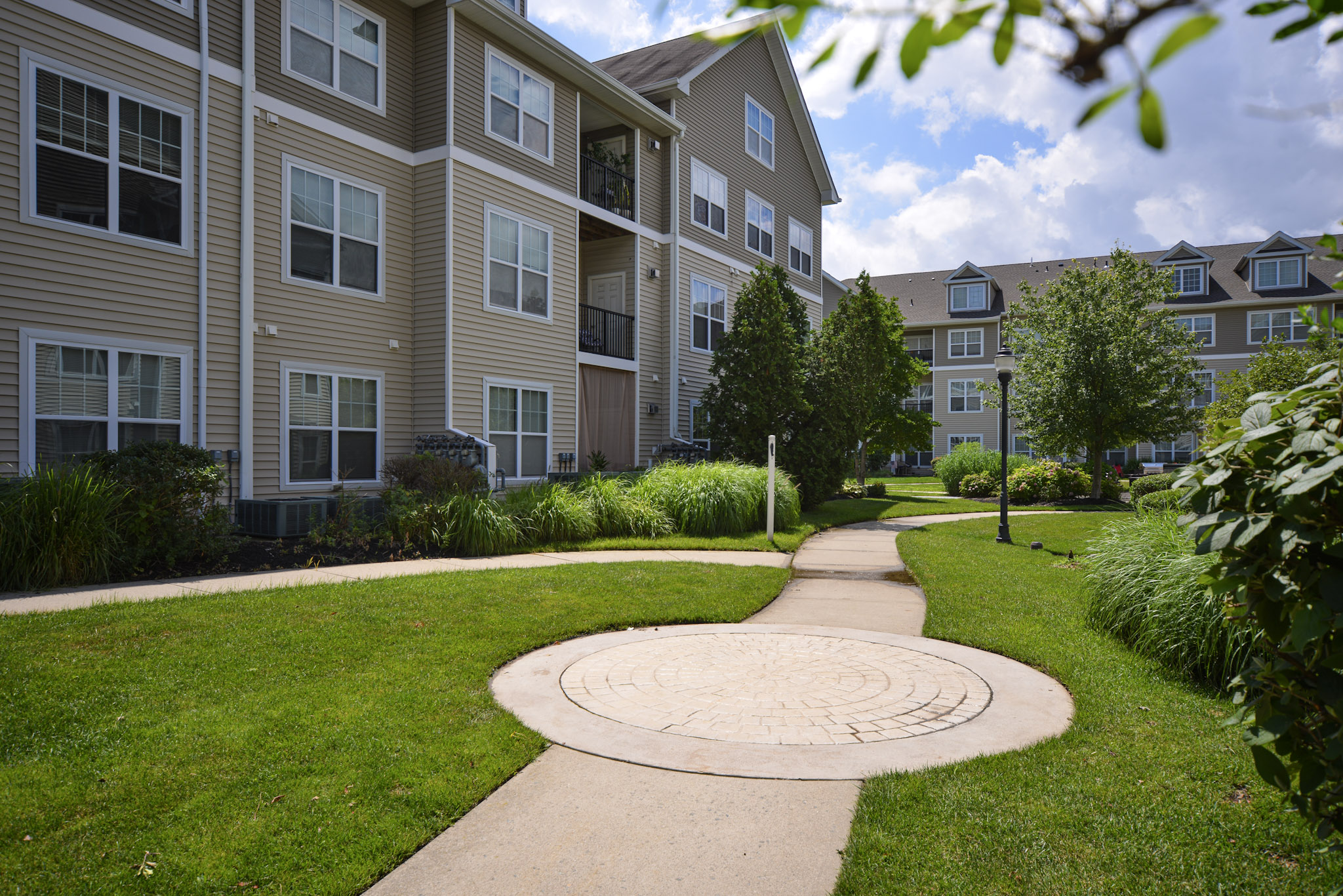 Pathway winding through a well-maintained green space with shrubs and small trees, leading up to a row of modern, light-colored multi-story apartment buildings under a partly cloudy sky. The area is inviting, featuring manicured lawns and a circular decorative concrete section.