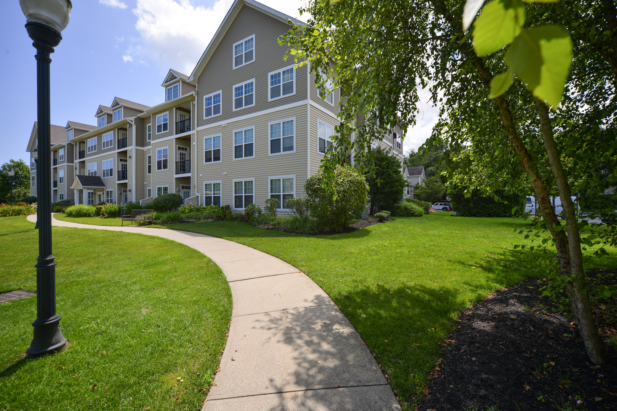 A well-maintained residential apartment complex surrounded by lush green lawns and landscaping. A paved pathway curves through the grass, leading towards the building. Trees and shrubs enhance the natural ambiance, with a lamp post visible along the walkway under a partly cloudy sky.