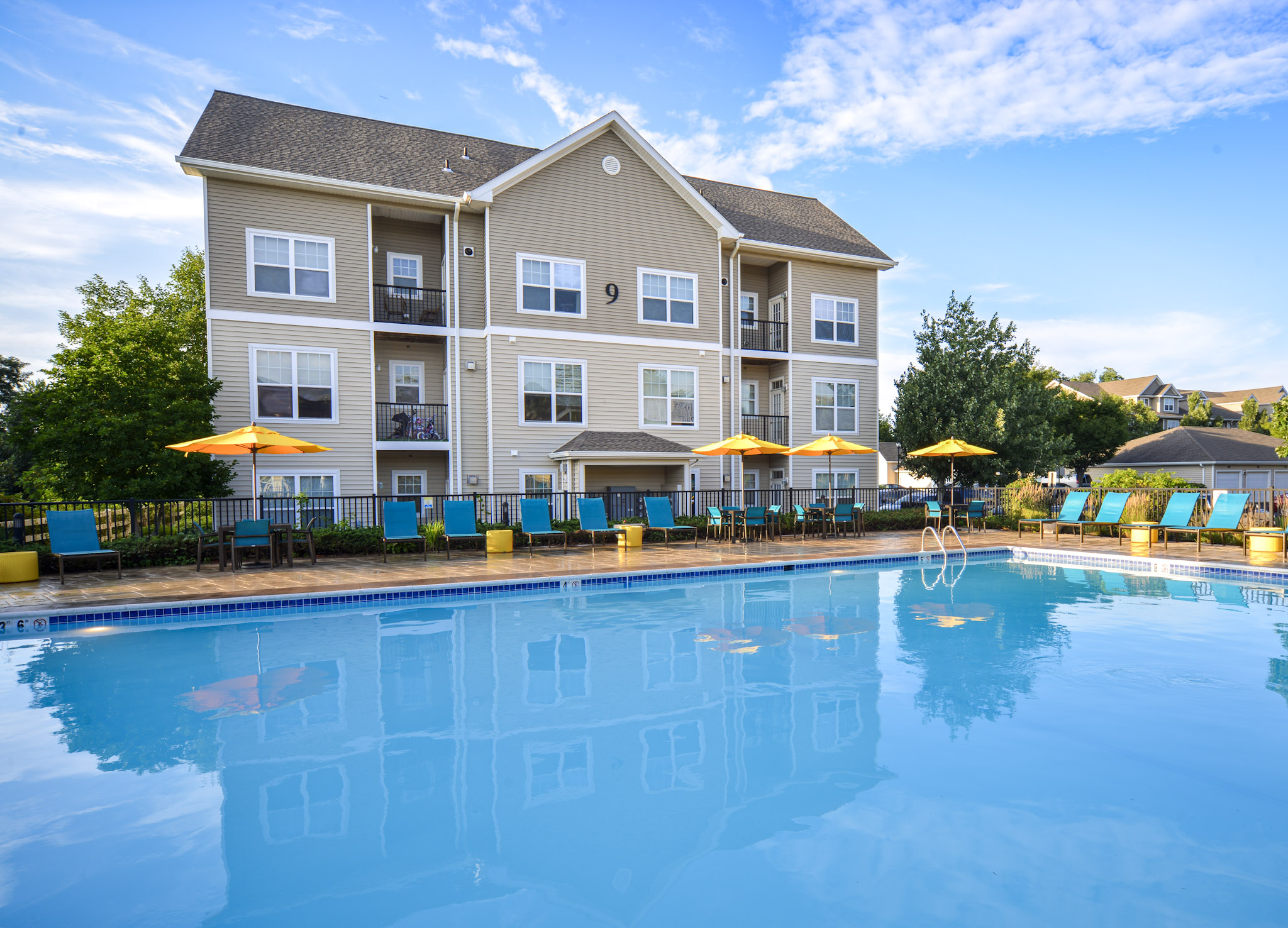 A well-maintained apartment complex featuring a large outdoor swimming pool surrounded by lounge chairs and umbrellas. The building has multiple balconies, greenery, and a clear blue sky above. Ideal for relaxation and leisure activities.