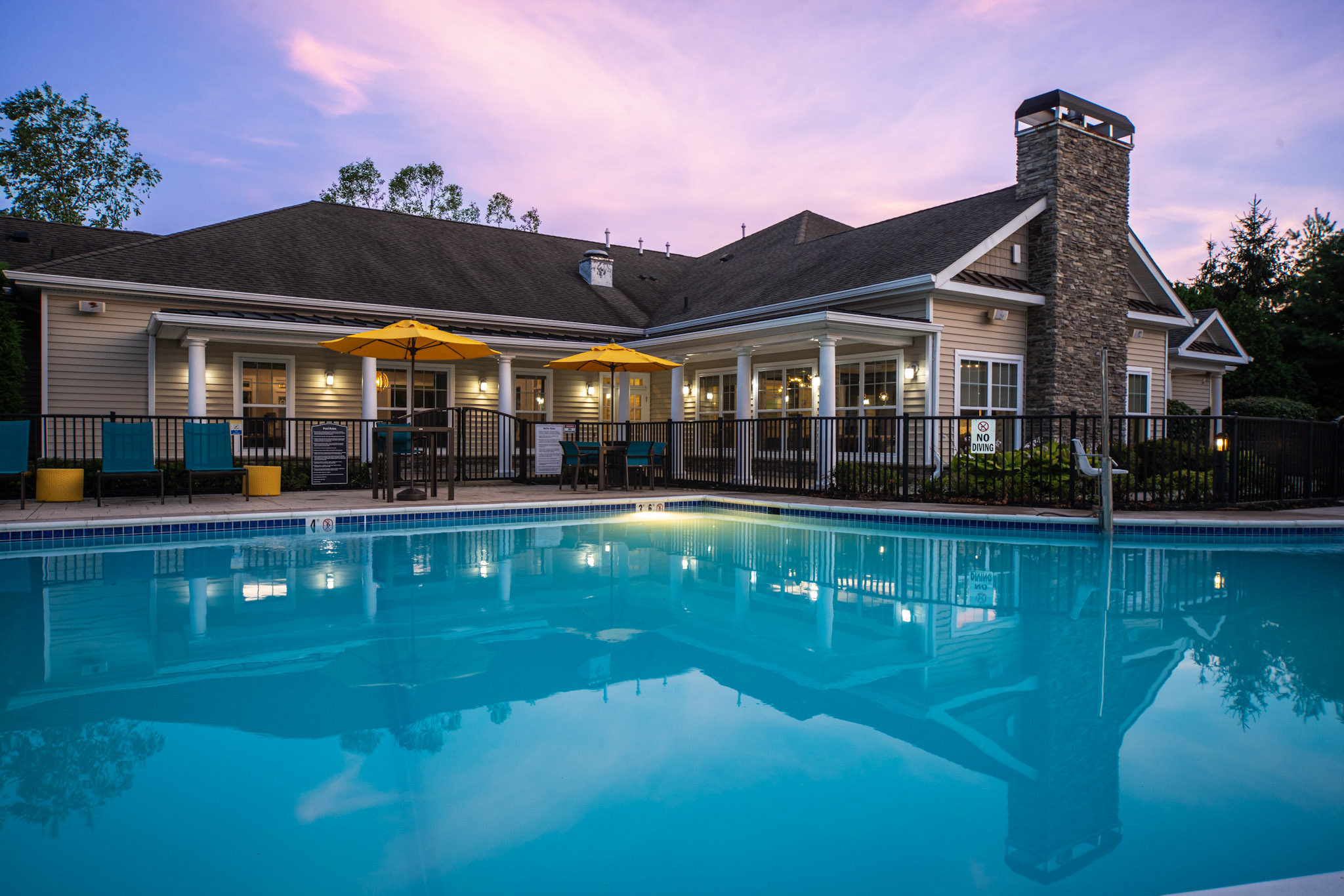 A serene swimming pool reflecting a sunset sky, surrounded by a well-maintained patio area. The pool features vibrant yellow umbrellas over seating areas, and a large building with glass windows and a stone chimney is visible in the background, adding to the inviting atmosphere.