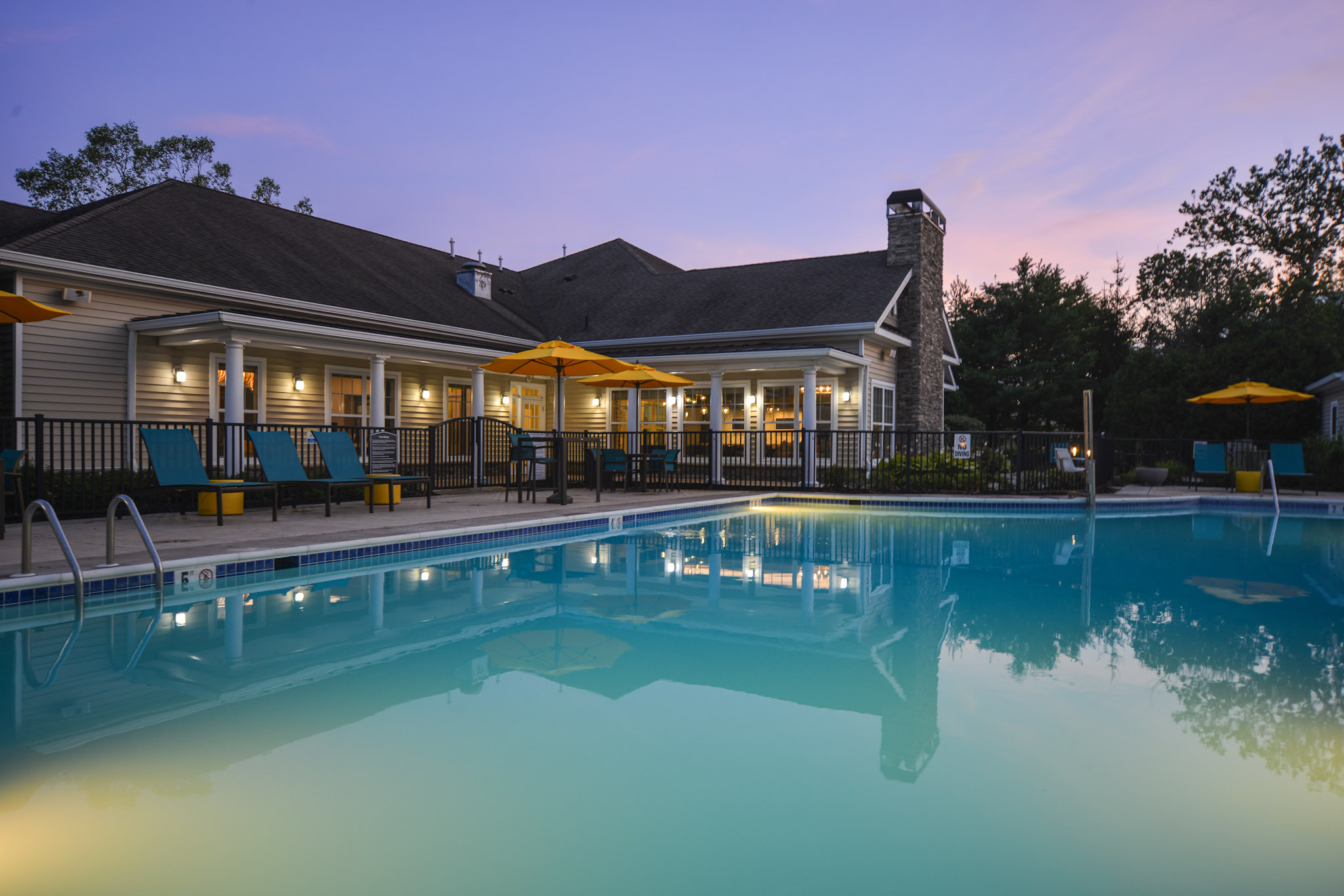 A tranquil outdoor pool area surrounded by lounge chairs and umbrellas, set against a backdrop of a modern building. The scene is illuminated by warm lights, with a colorful sunset sky reflecting in the clear water of the pool, creating a serene atmosphere.