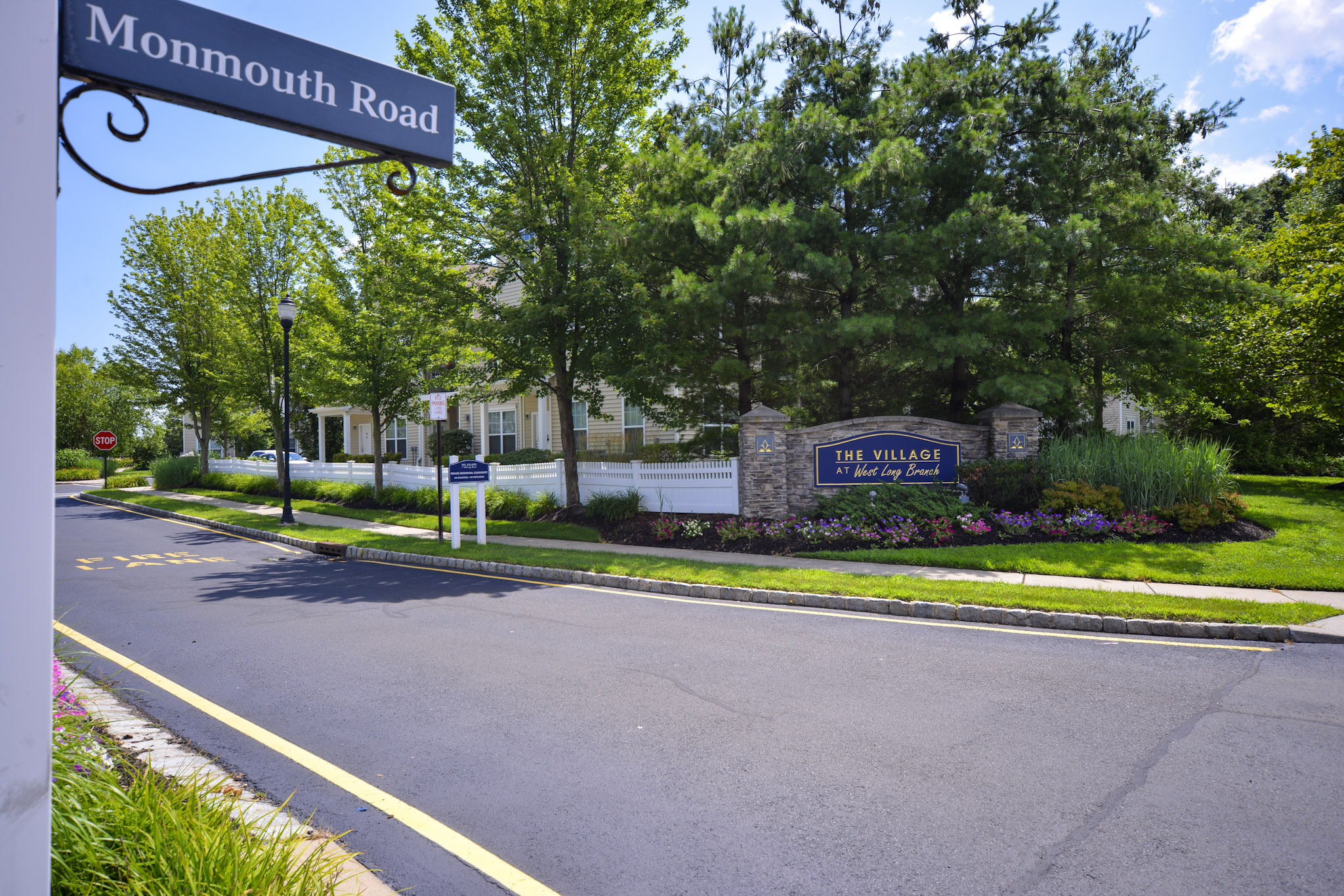 A scenic view of a residential area with a sign that reads "The Village at Long Branch." The entrance is marked by well-maintained landscaping, including trees and colorful flowers, along with street signs indicating "Monmouth Road" and a stop sign. The road is paved and leads into the community.