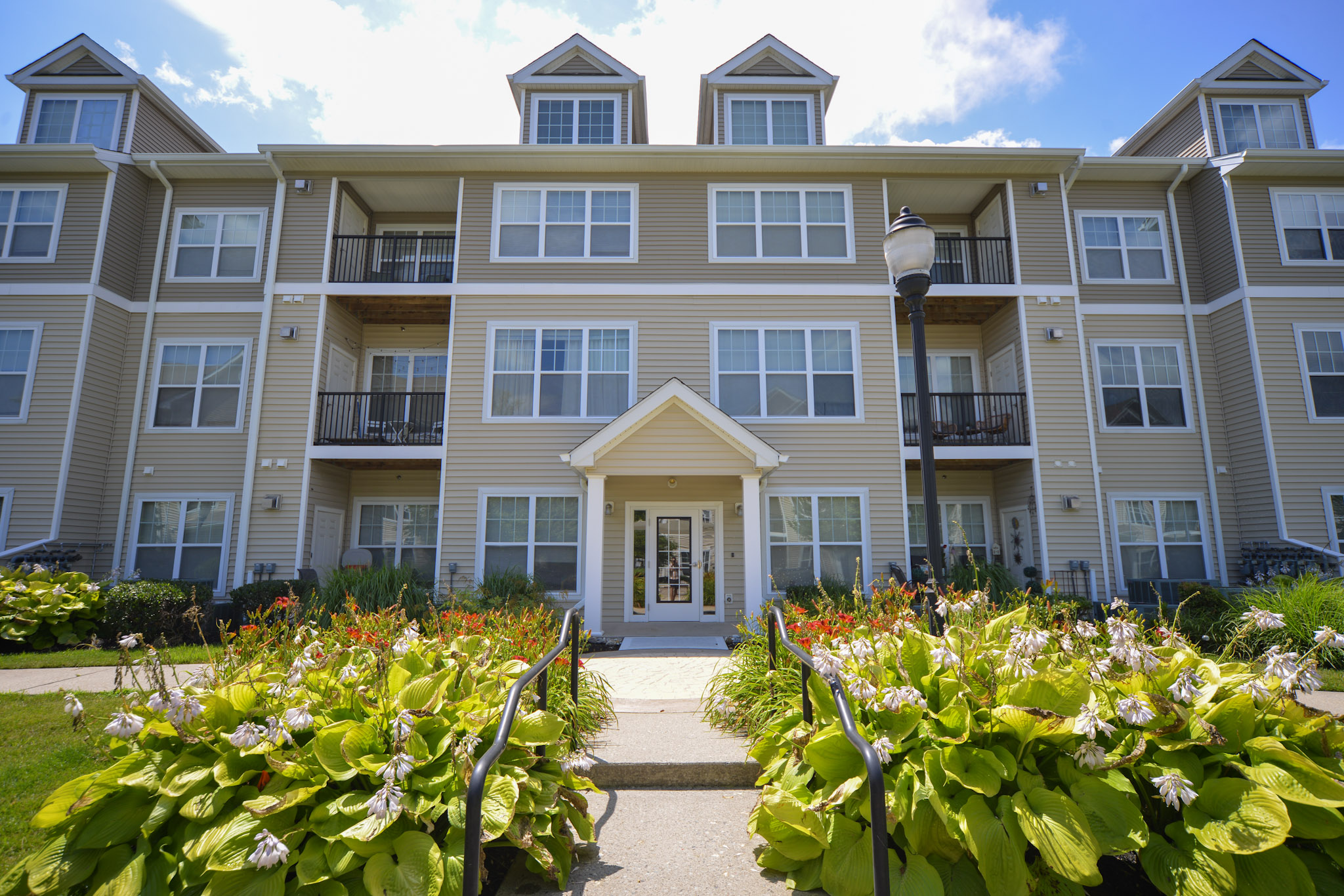 A three-story residential building with a beige exterior featuring multiple balconies. The entrance is framed by a small staircase and surrounded by well-maintained flower beds with lush green plants and colorful blooms, set against a clear blue sky.