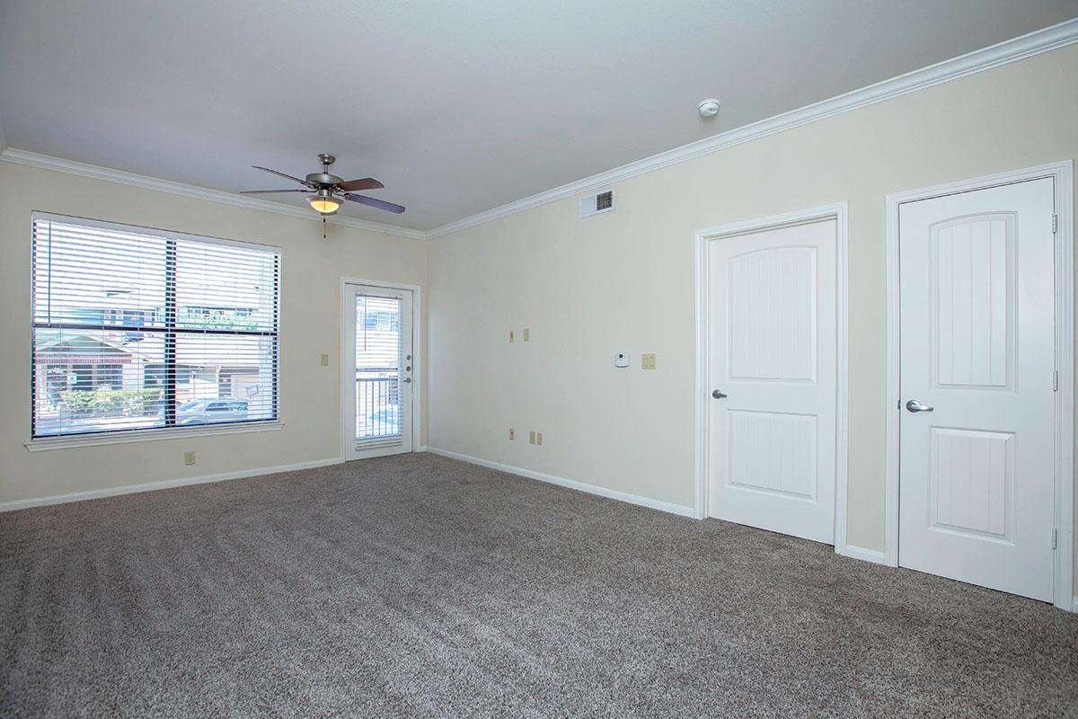A spacious, carpeted room with neutral-colored walls, featuring a ceiling fan, large window with blinds, and a door leading outside. Two closed doors are visible on the right side, and there are electrical outlets and wall switches. Natural light brightens the room, creating an inviting atmosphere.
