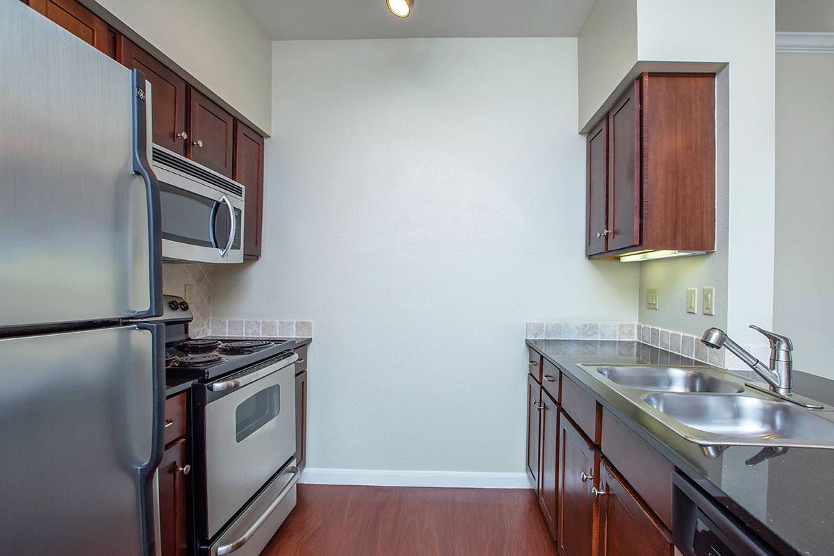 A modern kitchen featuring stainless steel appliances, including a refrigerator and stove. The cabinets have a dark finish, while the countertops are dark with a subtle texture. There is a double sink, and the walls are painted a light color, creating a bright and open atmosphere. Hardwood-like flooring completes the look.